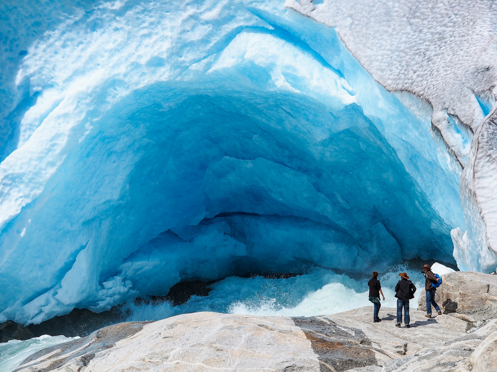 Three people stand looking into a blue-white ice cave, part of a glacier