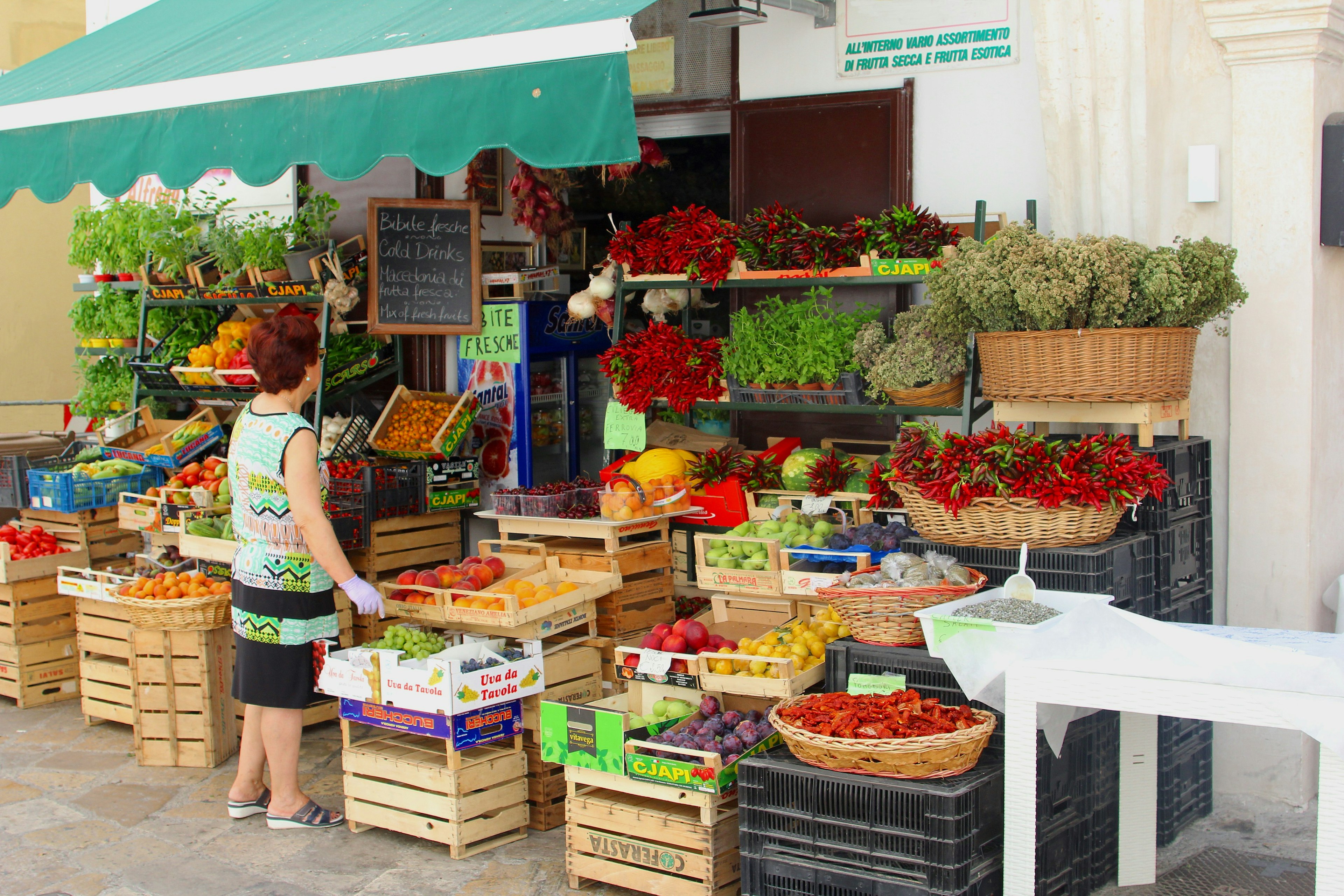 Italian woman with gloves buys fruits and vegetables in small grocery store, mini market with outdoor display of regional food products in neighborhood stree