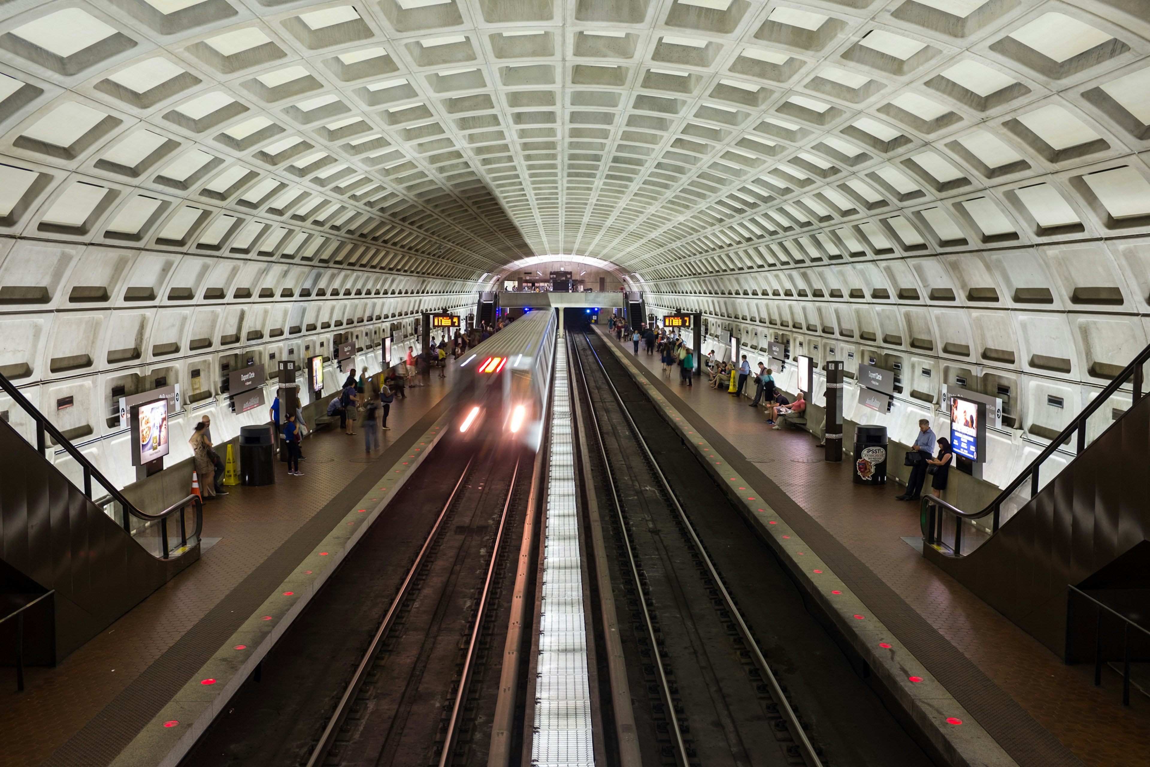 A metro train pulls into a station where people wait on the platform