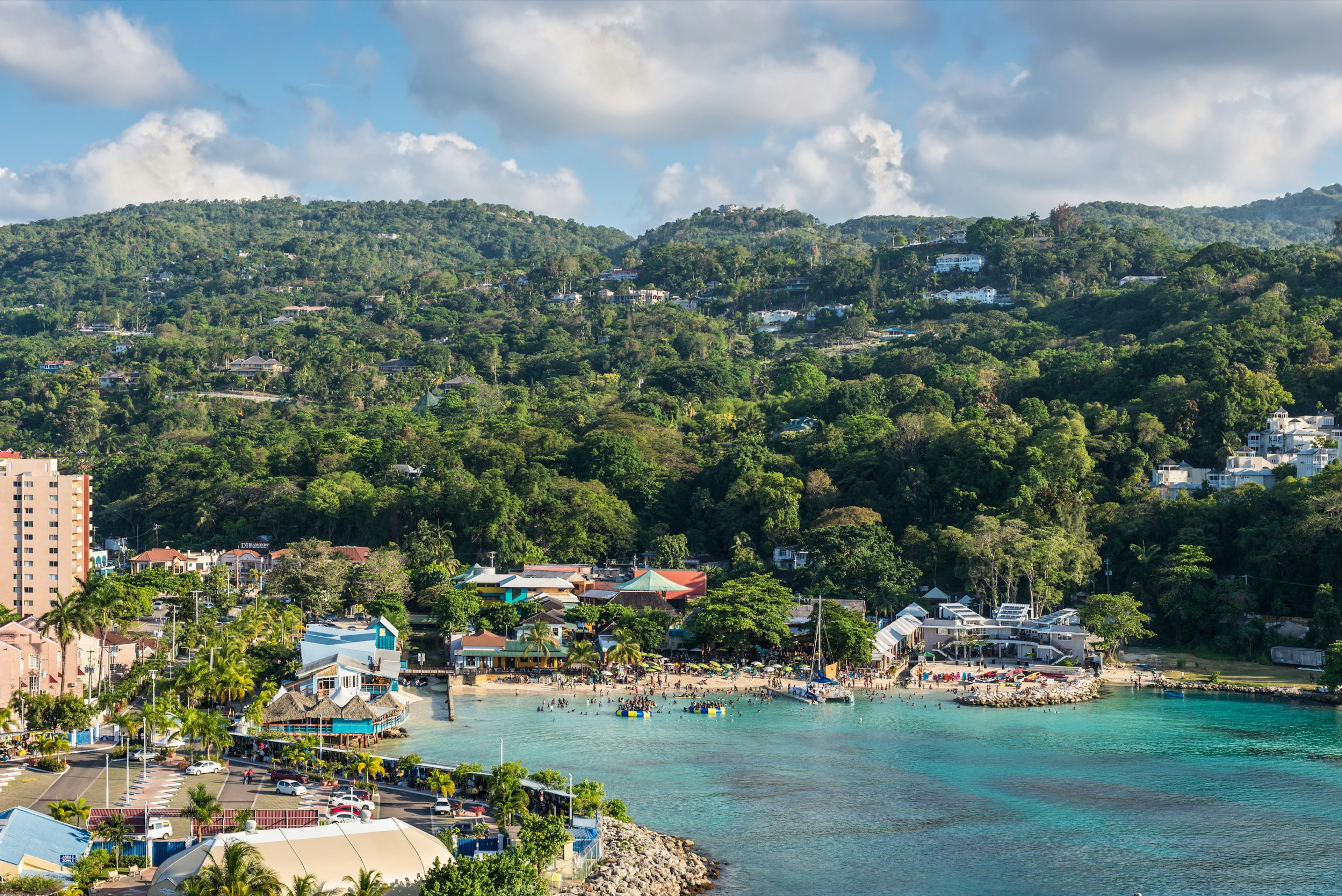 View from a ship of the blue waters of Fisherman's Beach in Ocho Rios, Jamaica. There are large groups of people are on the beach, there are large inflatable structures in the ocean and a sailboat docked in the shallow waters.