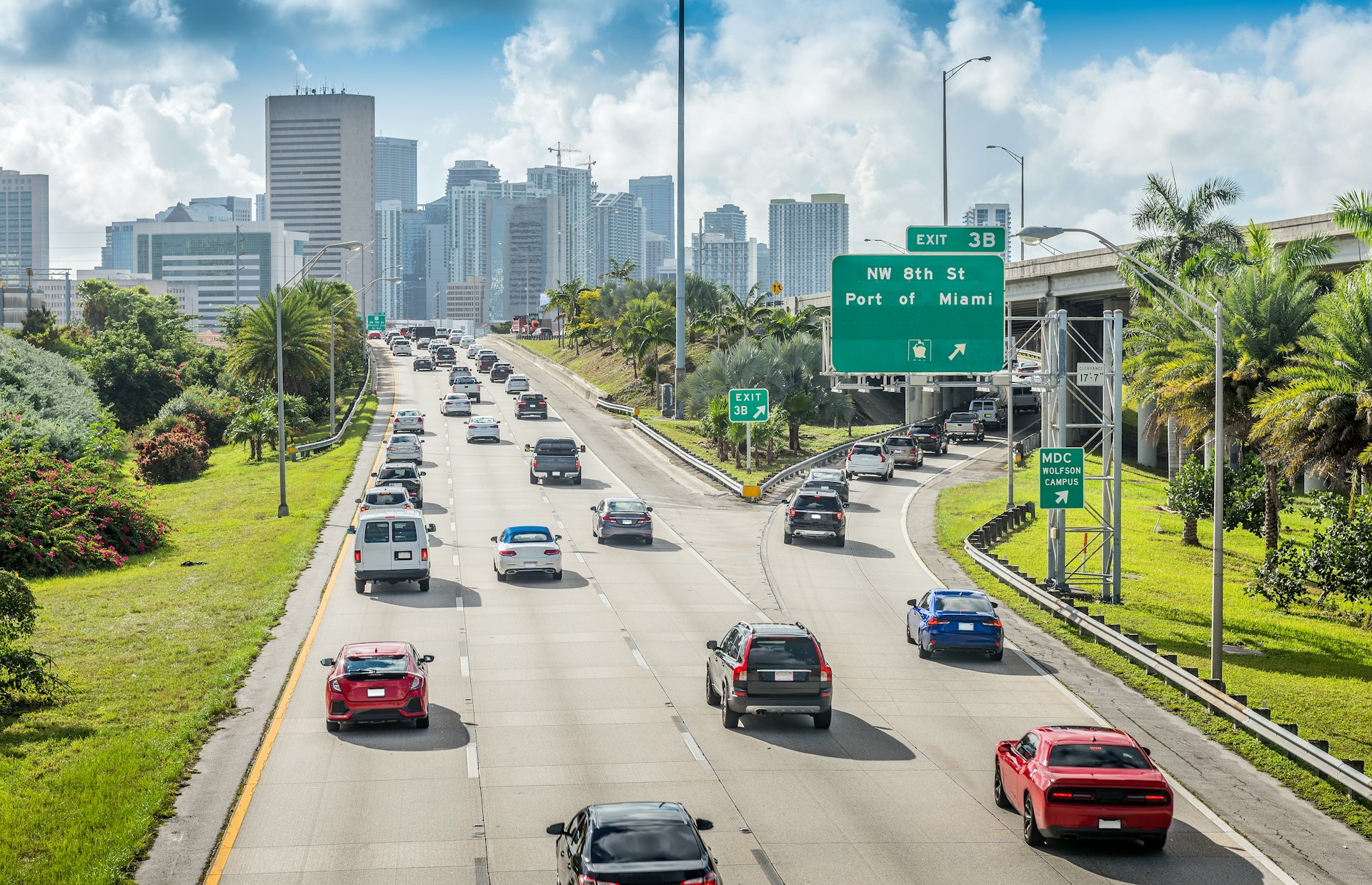 Many cars are driving on a three-lane expressway towards the skyscrapers of Miami Downtown during rush hour