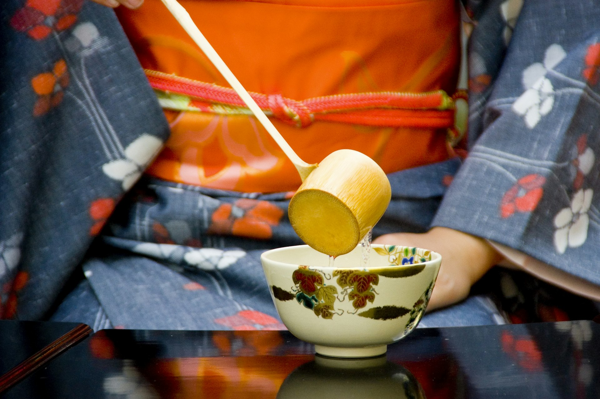 A horizontal image of a person conducting a tea ceremony, wearing a blue kimono and orange obi pouring hot water  from a wooden ladle into a Japanese tea cut sitting on a black lacquer table is shown