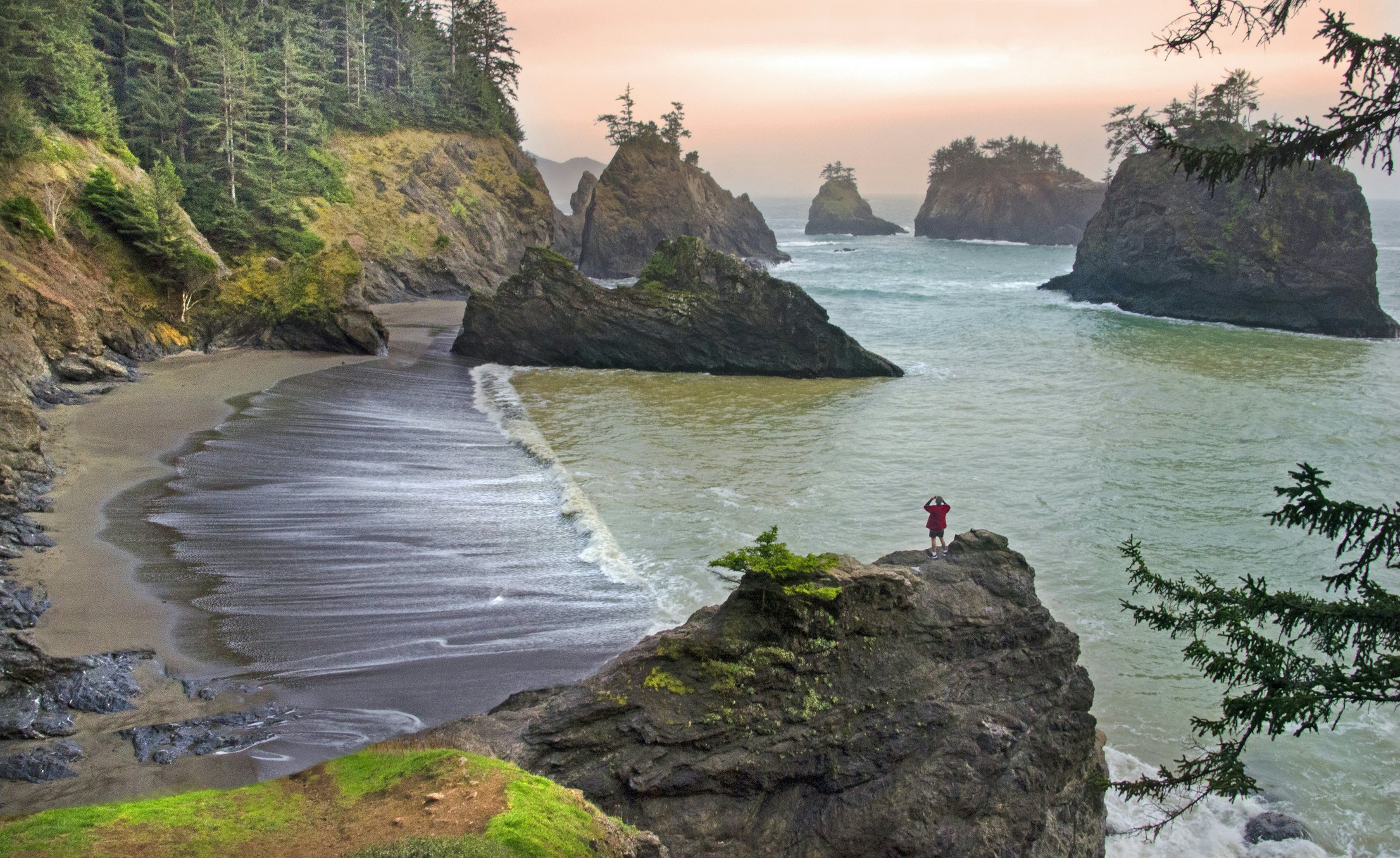 A person stands on a rock taking photos of a beach and rock stacks out at sea