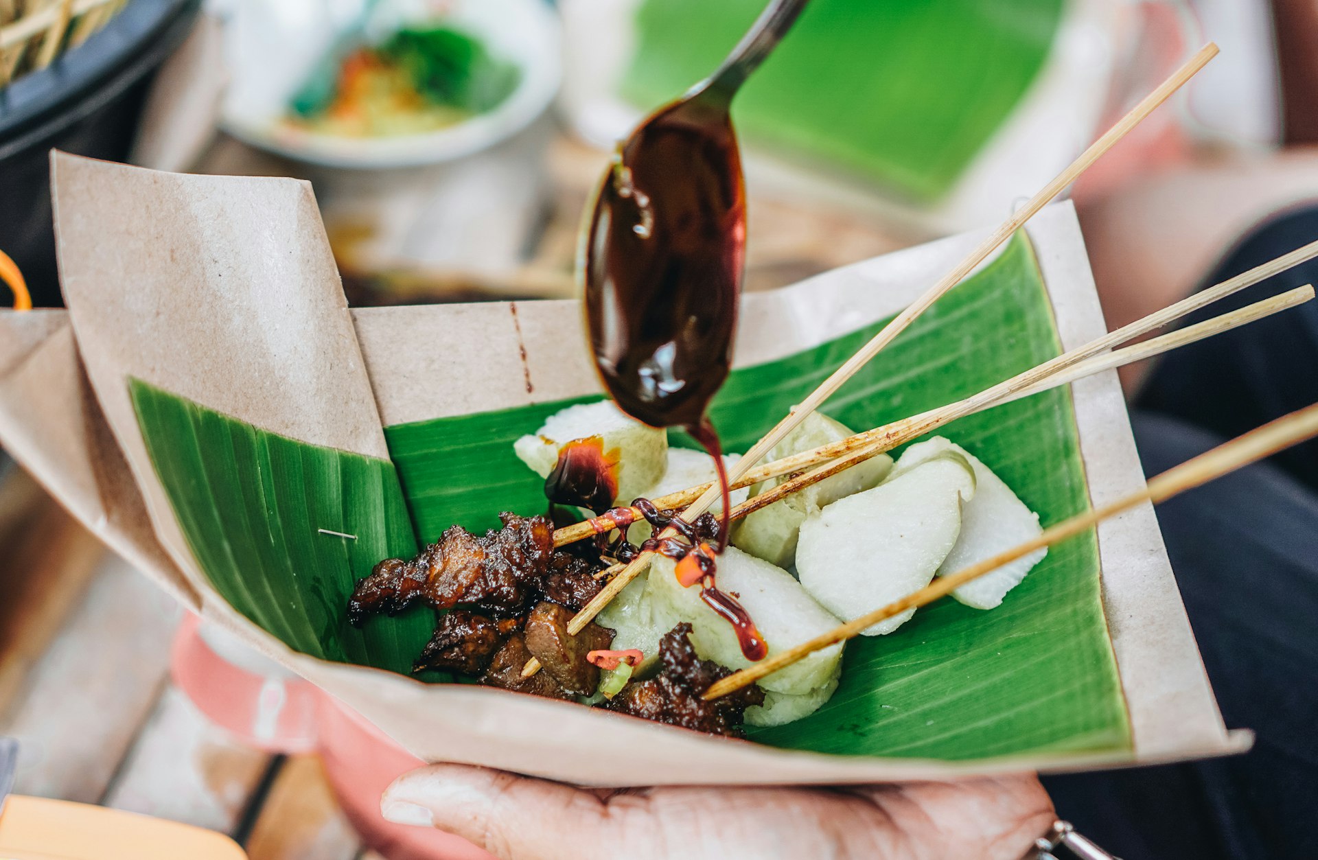 A banana leaf containing rice cake and meat skewers as a dark brown sauce drips onto the meat from a spoon