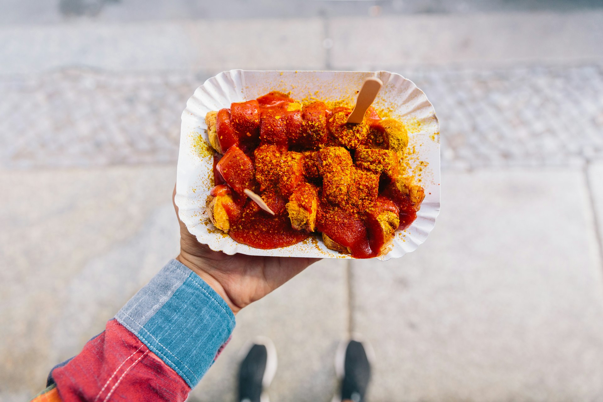 A hand holds out a small takeaway plate of curried sausage with a wooden fork