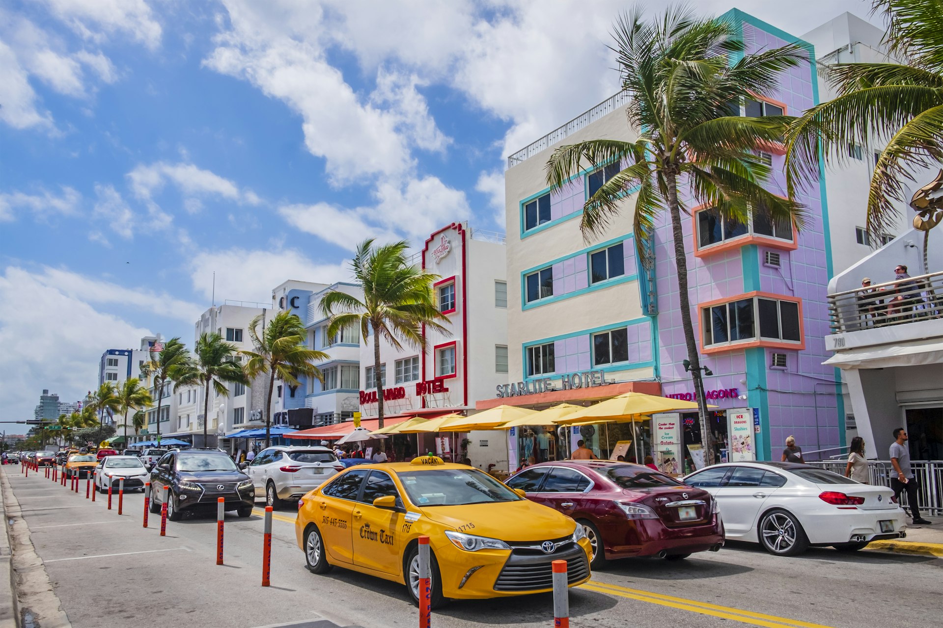 A bright yellow taxi is at the front of a line of cars driving along Ocean Drive in Miami, where tall palm trees stand in front of art deco buildings