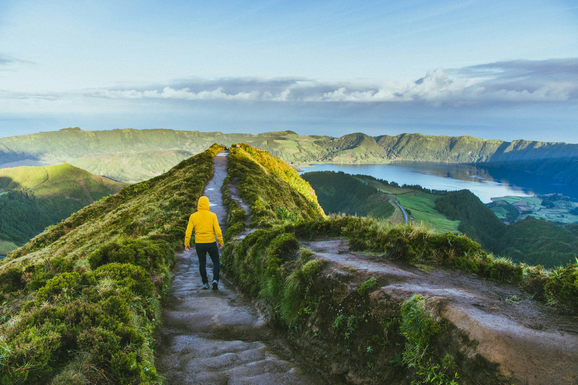 A person in a yellow rain jacket walks down a path between two craters filled with lagoons