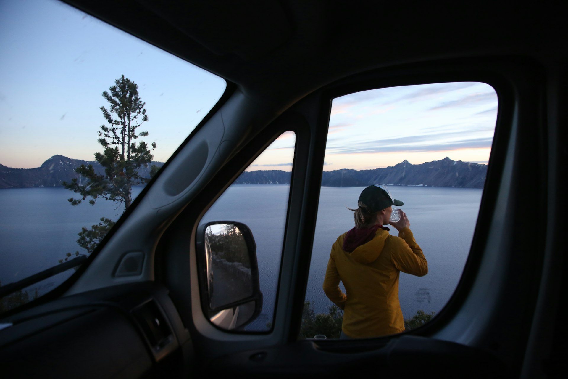 A young woman enjoys a drink by her car on the edge of Crater Lake, Crater Lake National Park, Oregon, USA
