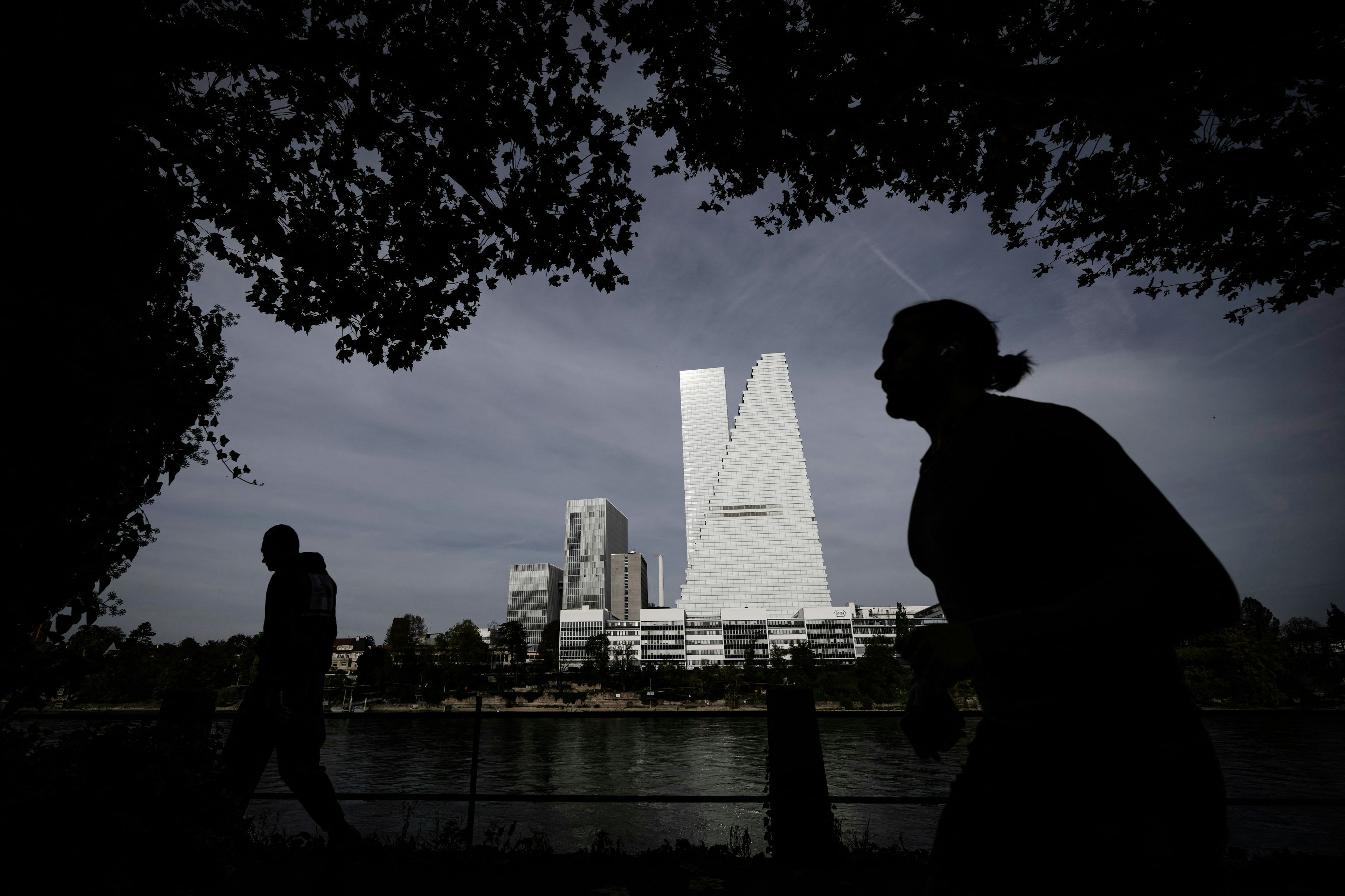 Passers-by are silhouetted as they walk along the banks of the River Rhine next to the Roche Towers, Basel, Switzerland