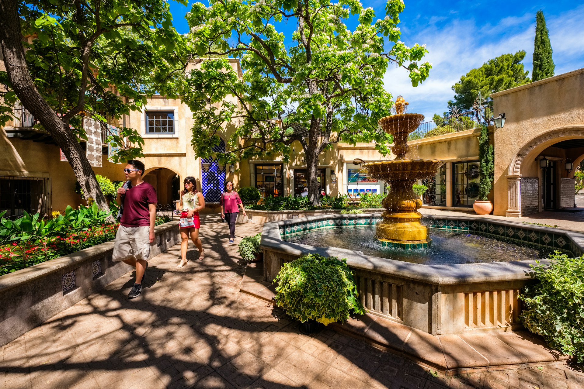 Three people walk alongside a fountain in an arts and shopping complex, shaded by trees