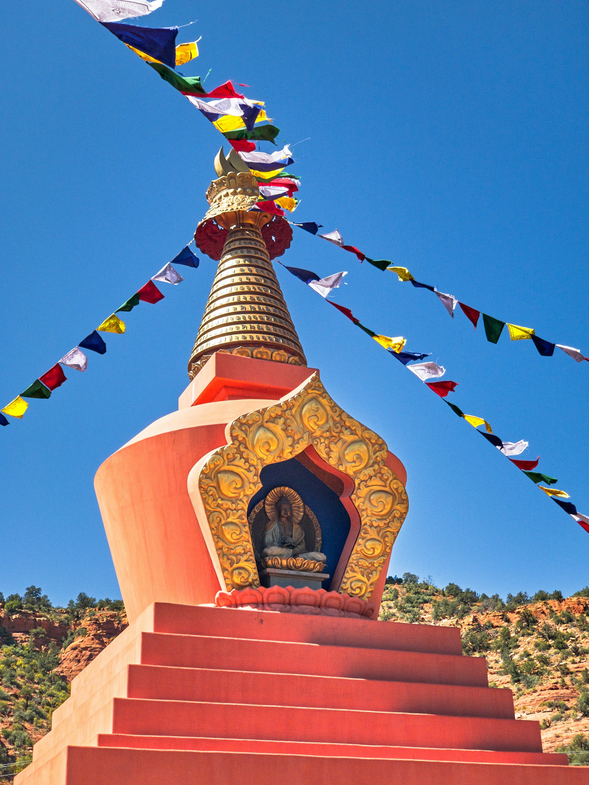 A terracotta-colored Buddhist stupa set against a clear blue sky, with colorful prayer flags strung from the top.