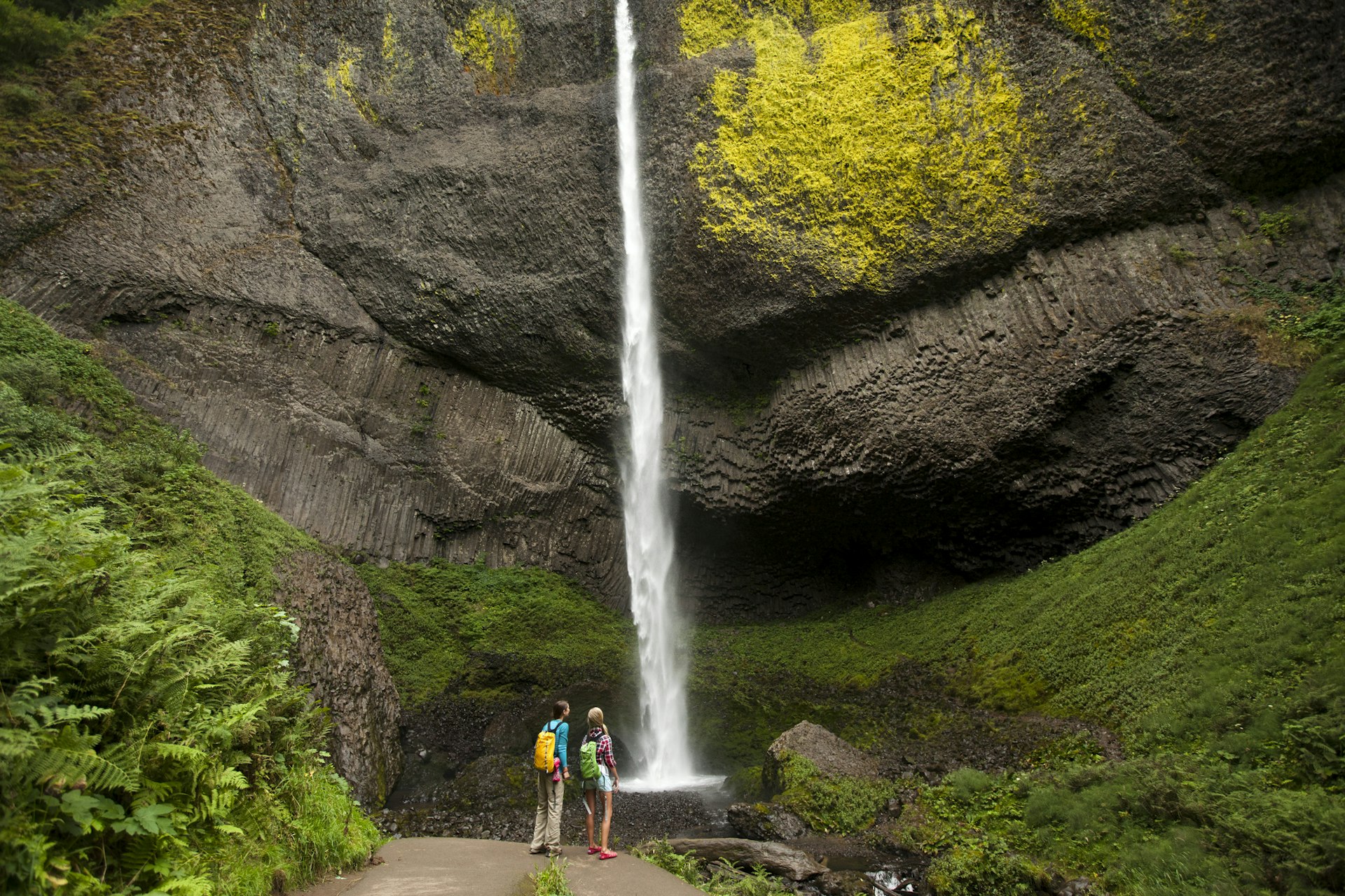 Two hikers stand at the base of a waterfall looking upwards