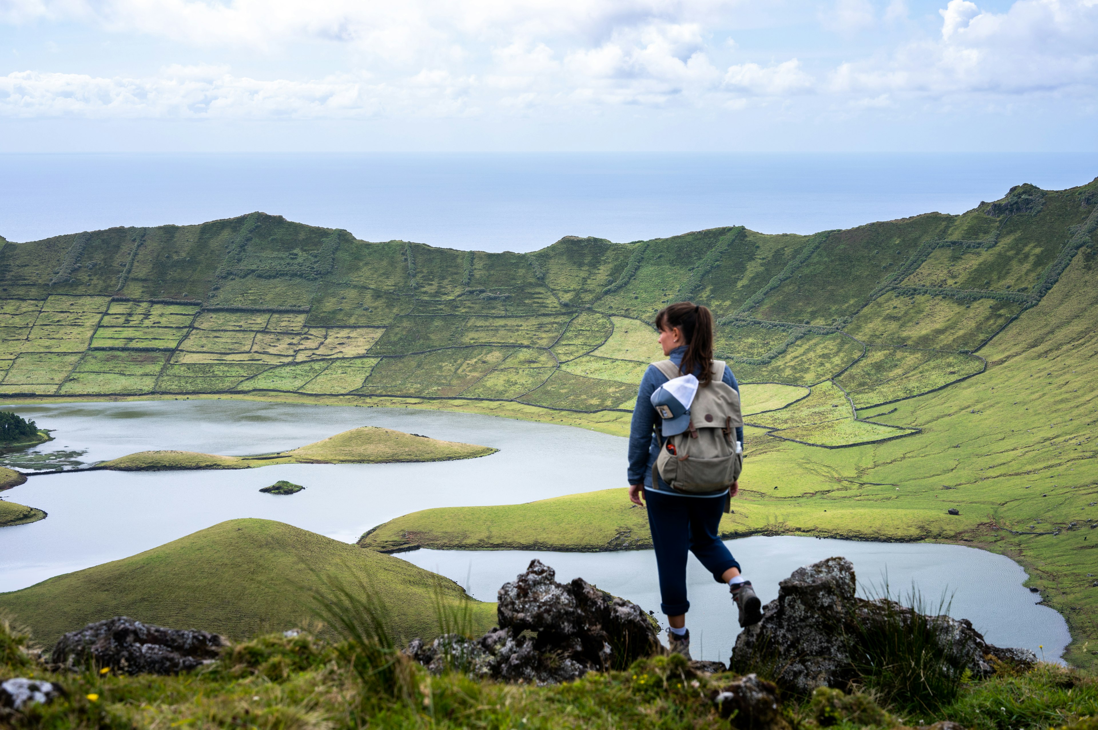 A woman stands in front of a crater covered in green, with a lake at the bottom