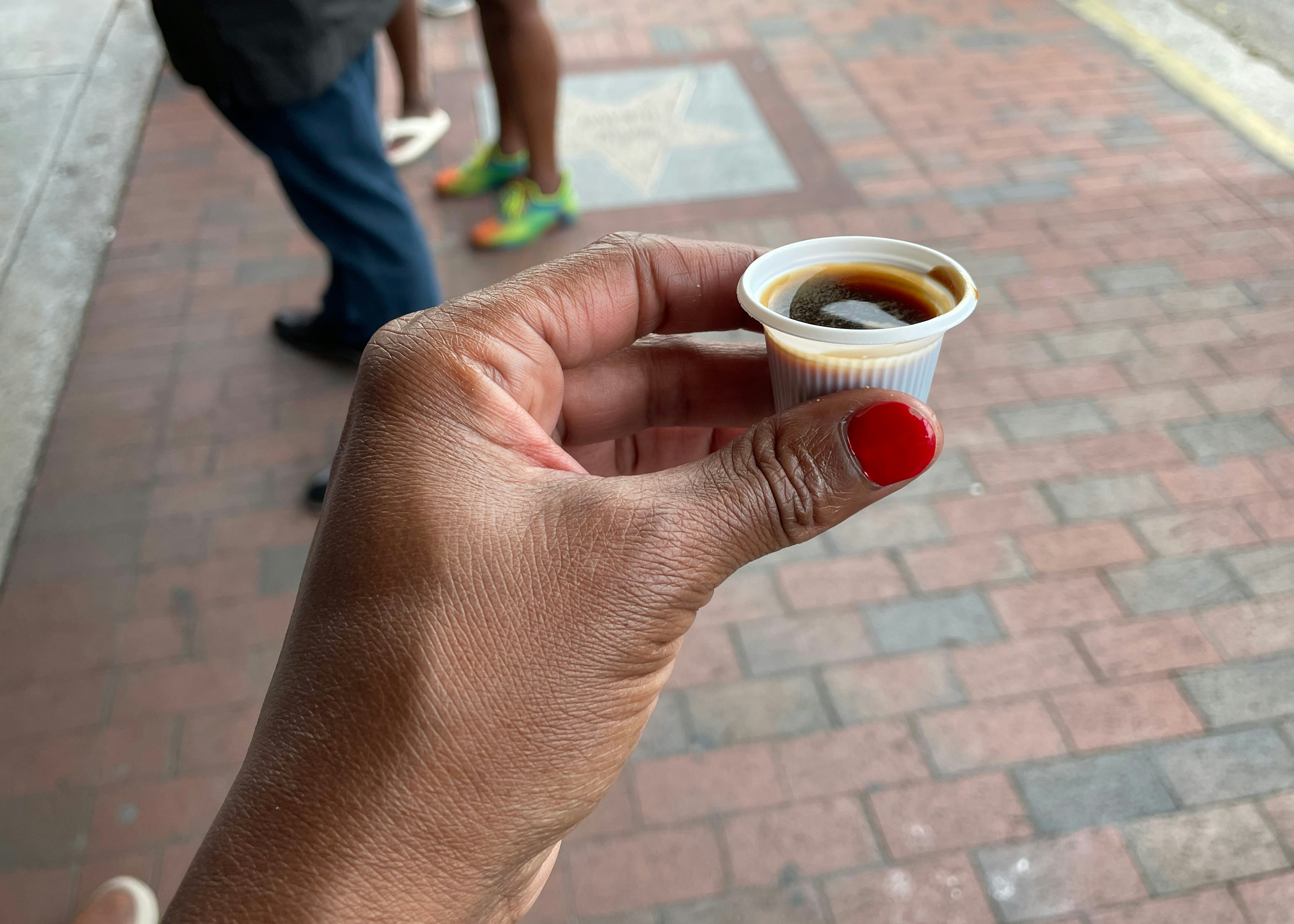 A hand is holding a thimble-sized plastic cup of black coffee