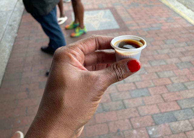 A hand is holding a thimble-sized plastic cup of black coffee