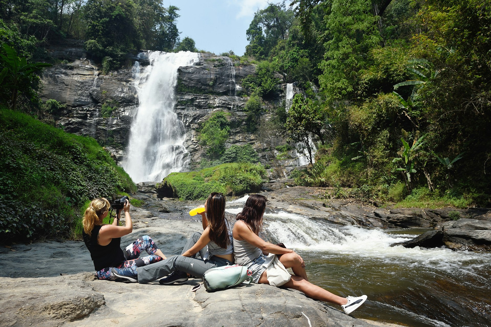 Three people sit on a rock near the pool of a fast-flowing waterfall 
