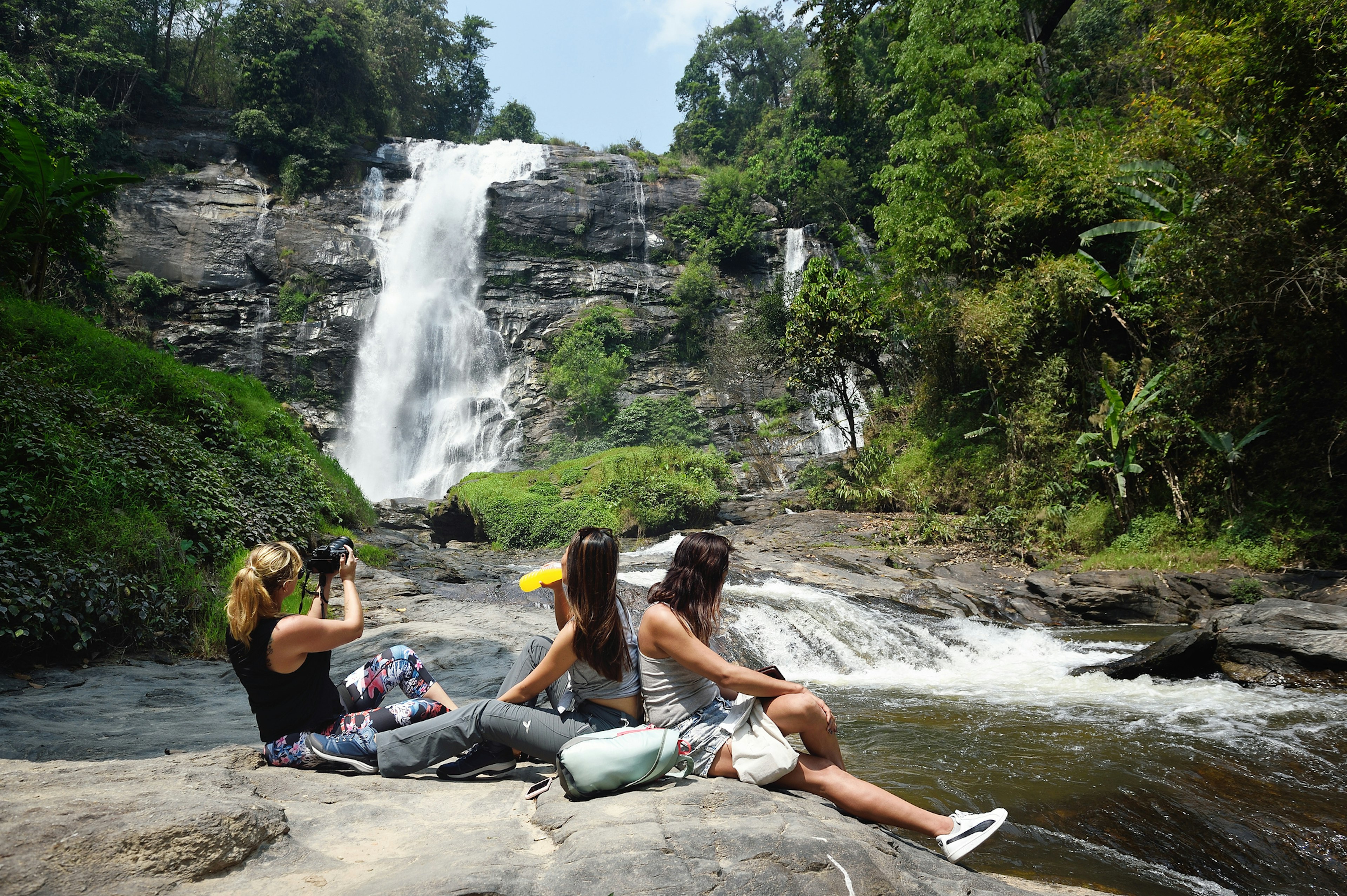 Three people sit on a rock near the pool of a fast-flowing waterfall