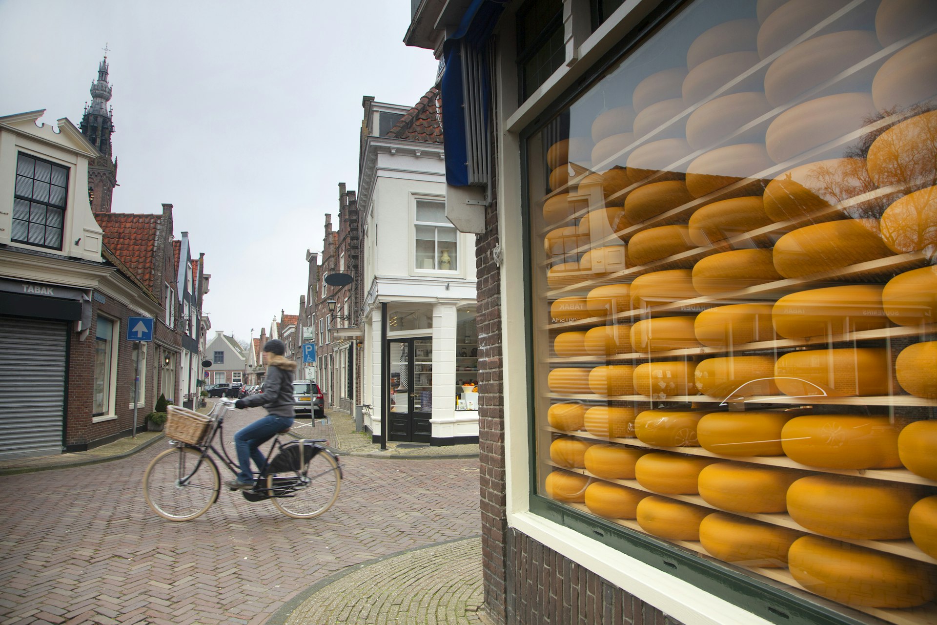 Netherlands, Edam town, female , young lady on bicycle riding near shops. on right of image a cheese shop display.