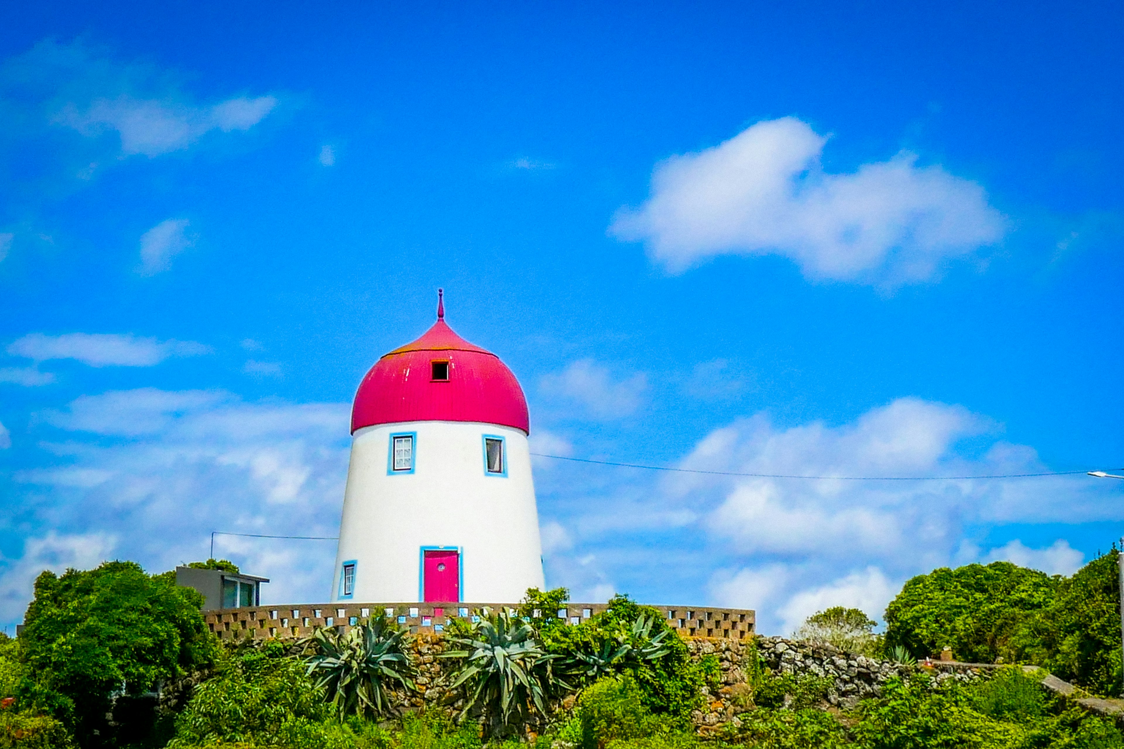 A red-roofed, Dutch-style windmill without its sails stands against a bright blue sky.