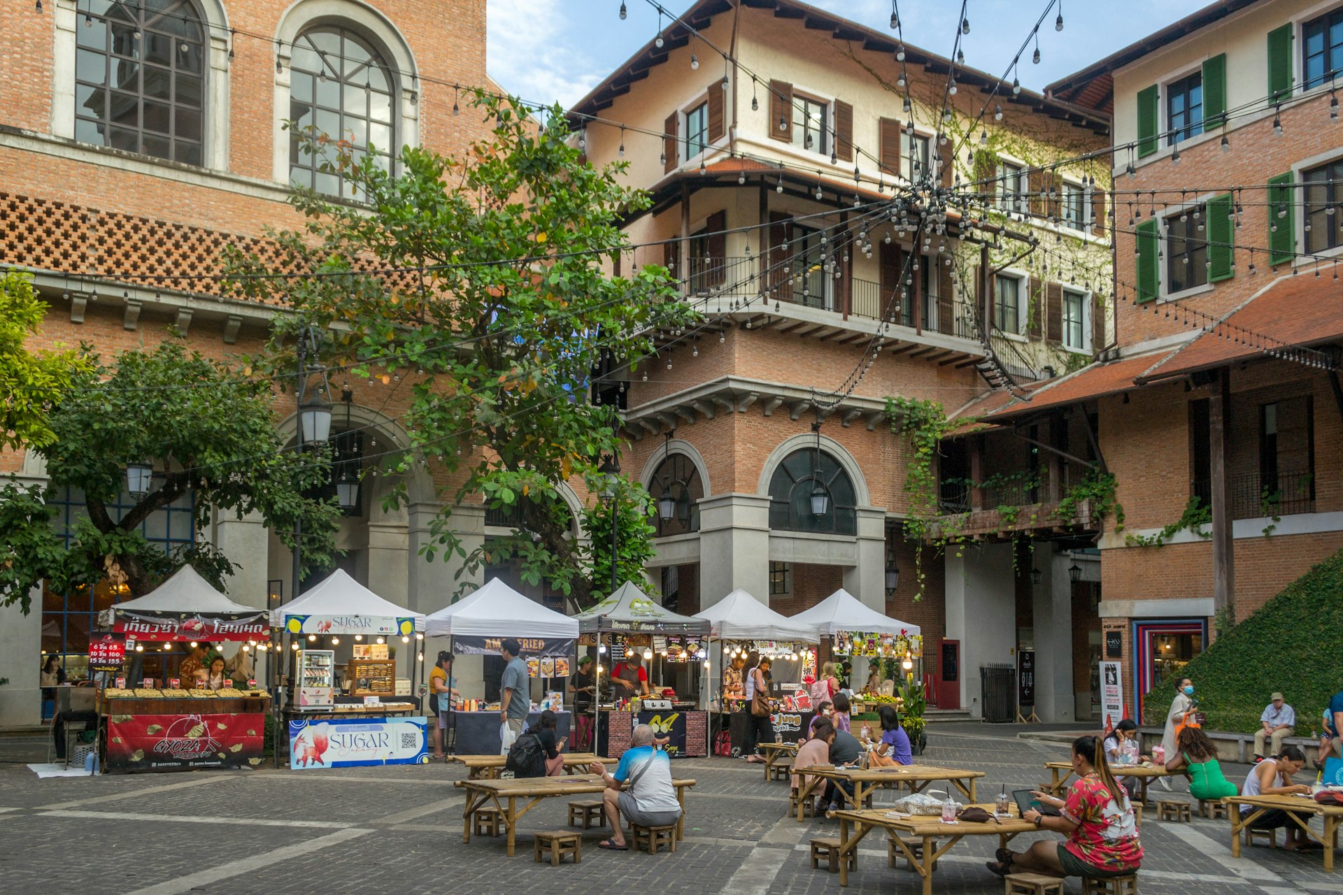 A handful of street food stalls are set up in the courtyard of the One Nimman retail complex, and people are sitting at picnic tables and eating. 