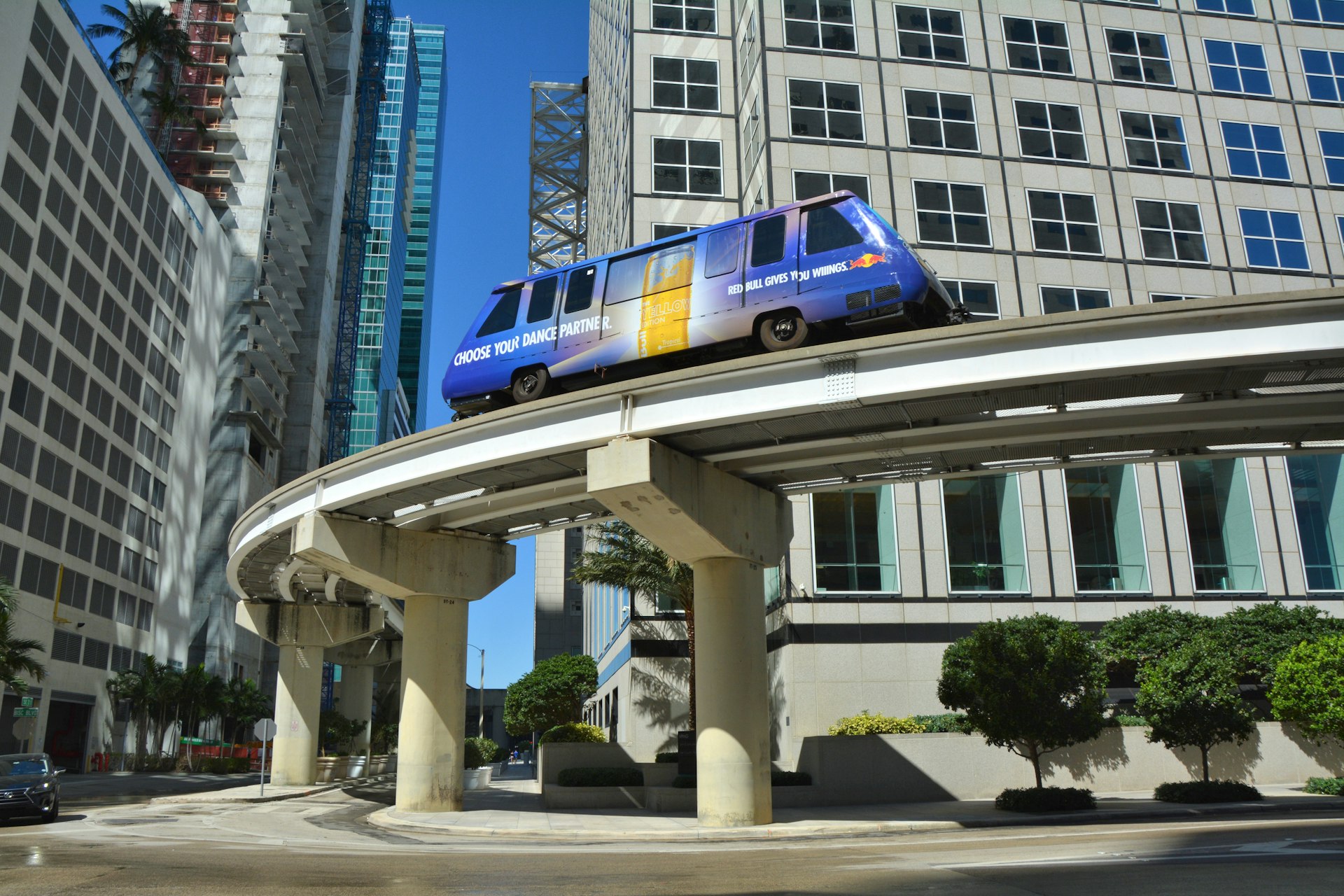 A one-car Metromover monorail train moves along an elevated track between high-rise buildings in Miami