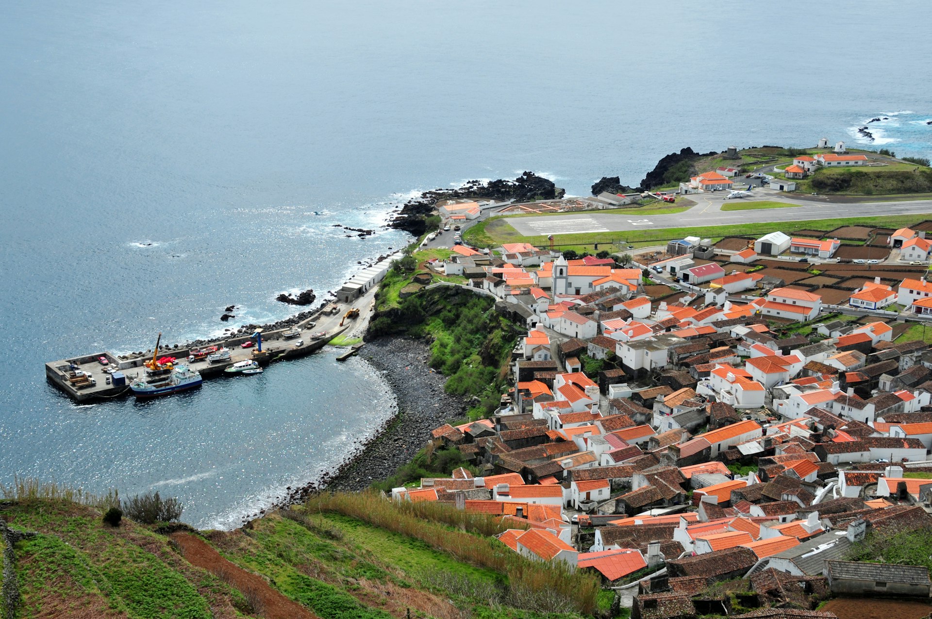 Vila Nova do Corvo, Corvo Island, Azores, Portugal: red roofs of the island's main settlement seen from the hills - view over the town with the volcanic rock beach, boats docked in the harbor's pier