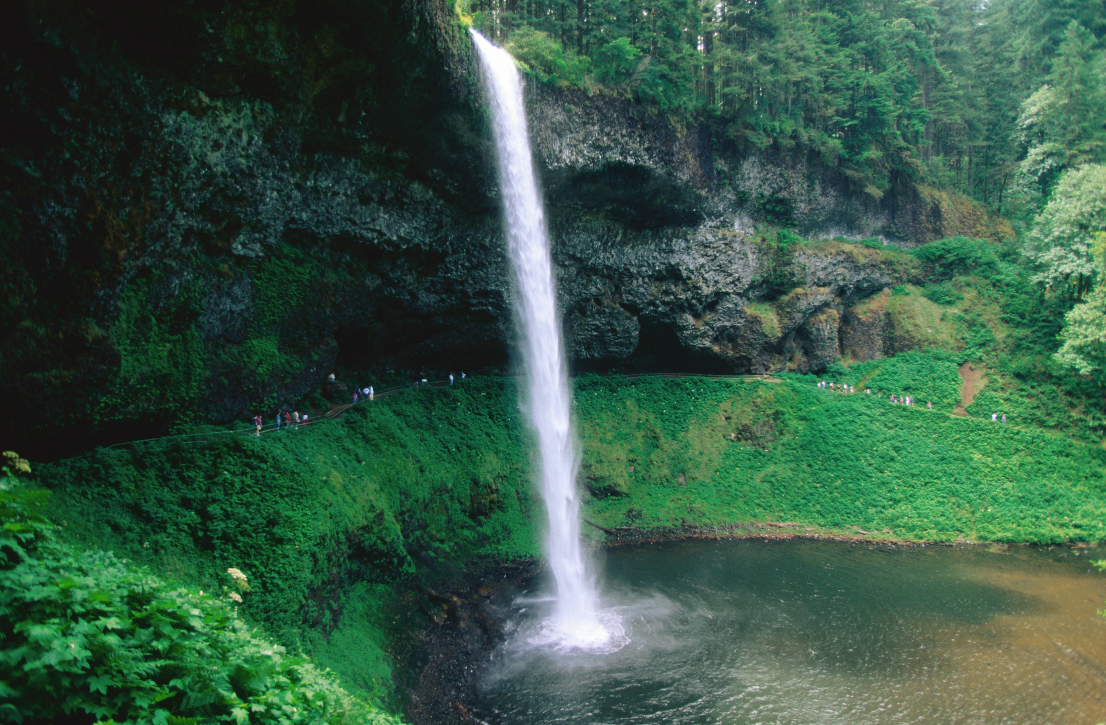 A cascading waterfall plunges into a pool. People on the hiking trail behind it are like tiny specks in comparison