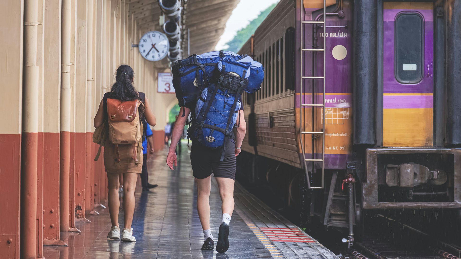 A man and a woman wearing backpacks walk towards a train that's waiting on the platform at Chiang Mai train station.