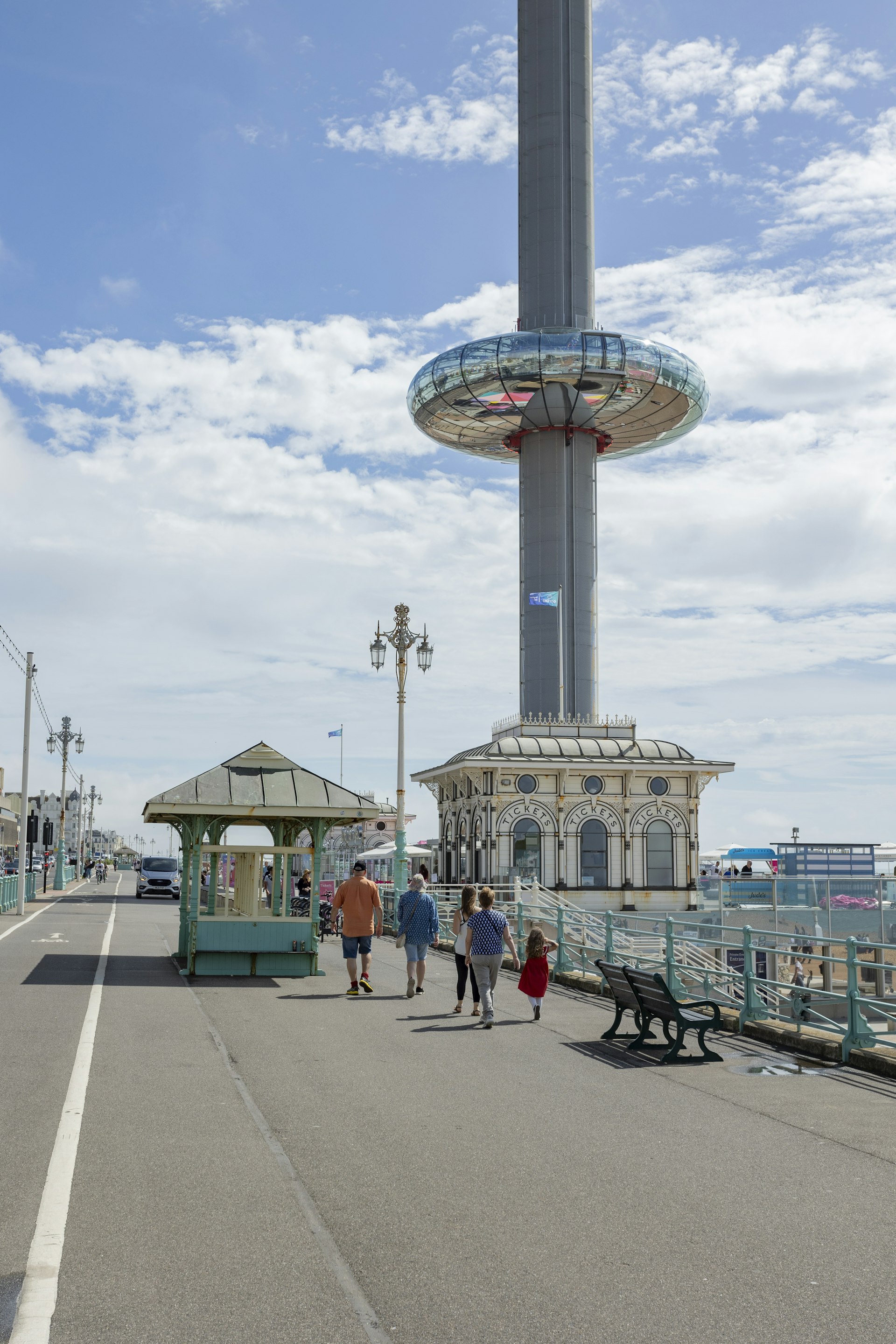 A circular glass observation pod is ascending up a high tower on a beach promenade