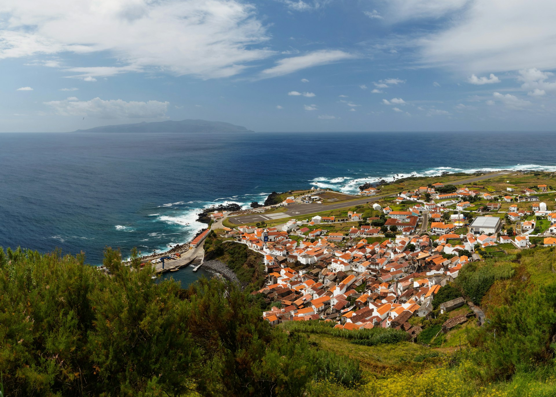 A view looking down on Vila do Corvo, a small town with tightly packed houses with terracotta rooftops.The island of Flores is visible on the horizon.