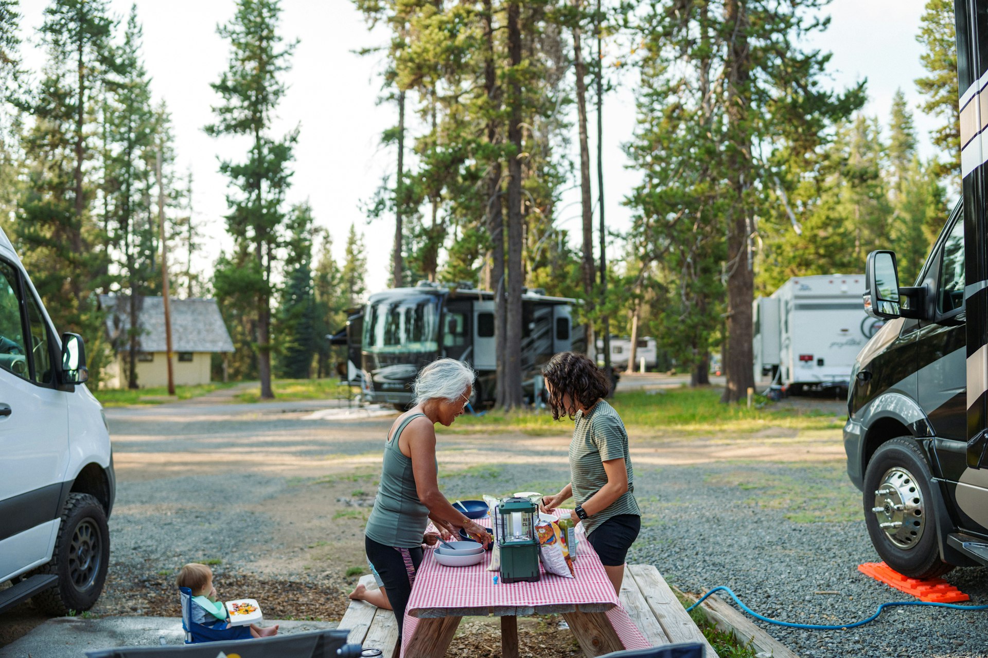 Two women prepare a meal at a picnic table beside an RV camper parked in a scenic tree-lined campsite