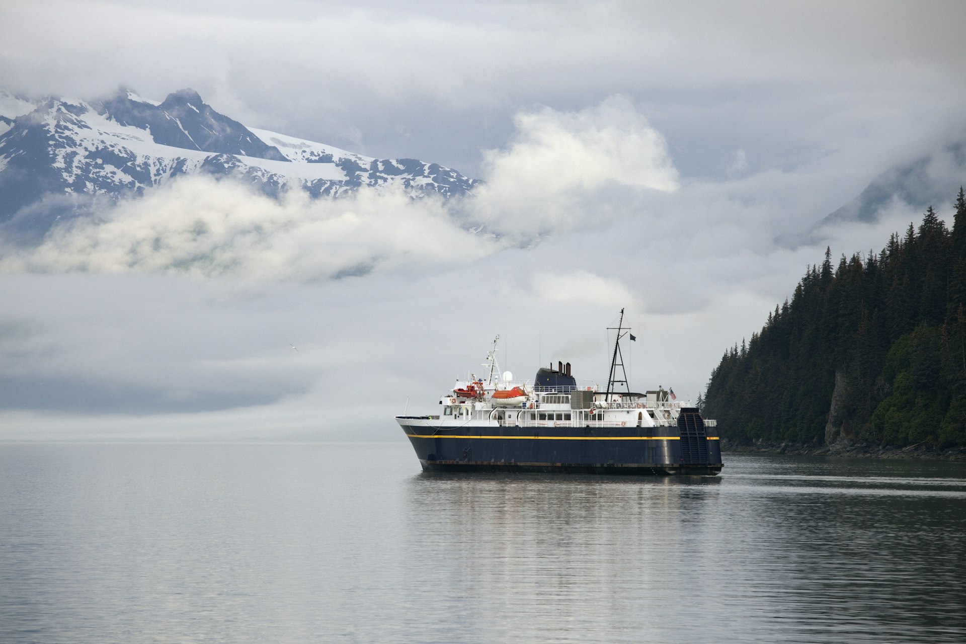 A large ferry leaving a port backed by mountains covered with cloud