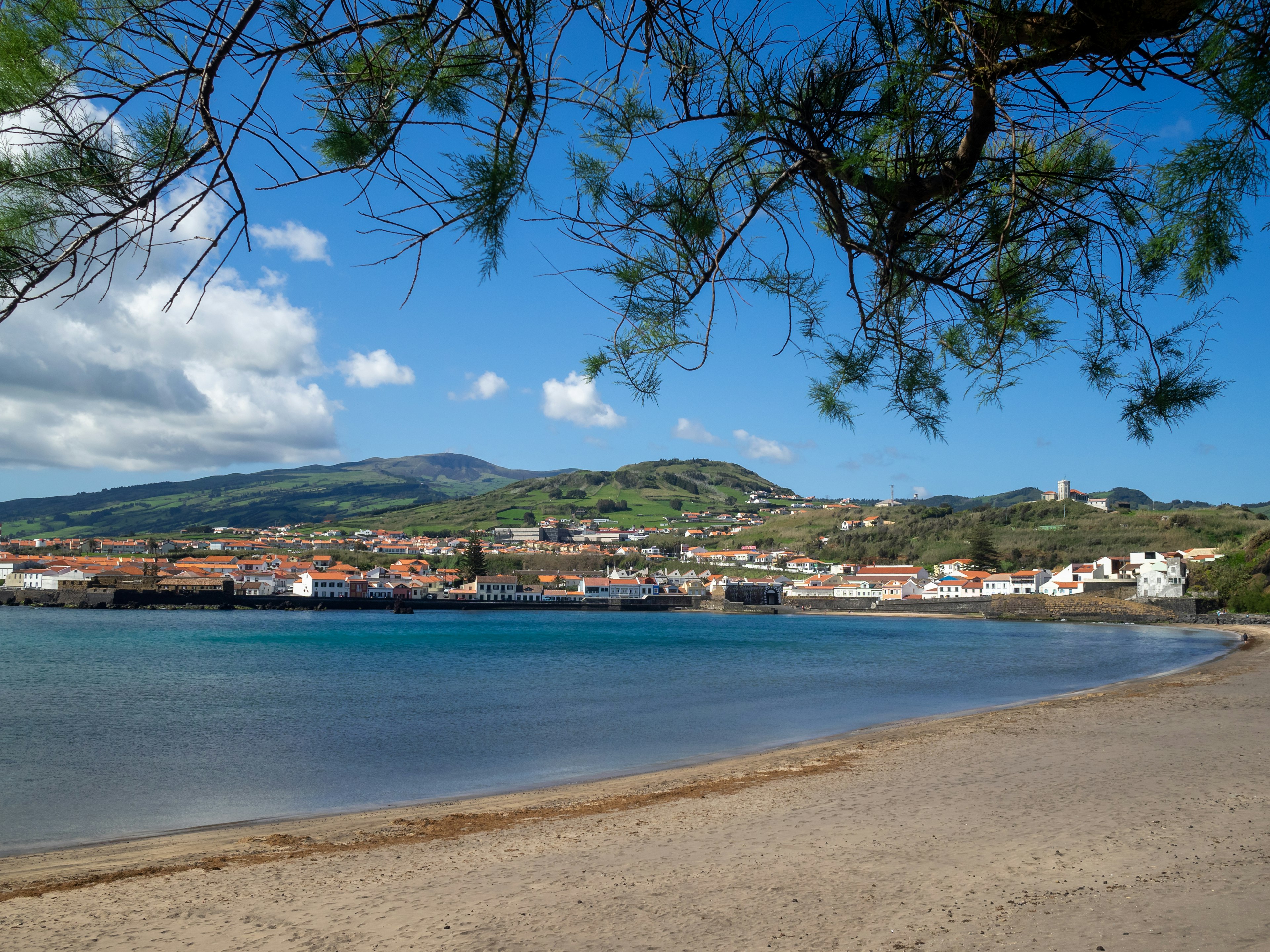 A gently curving stretch of light brown sand leading to calm water, with a town visible on the other side of the bay.