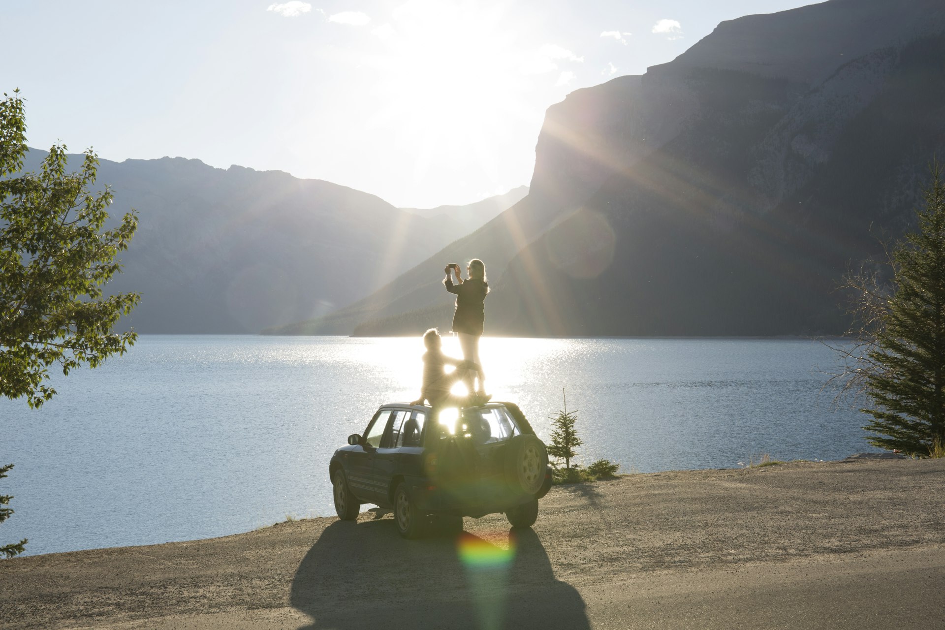Woman takes photo from car roof while man relaxes by a lake, Banff National Park, Alberta, Canada