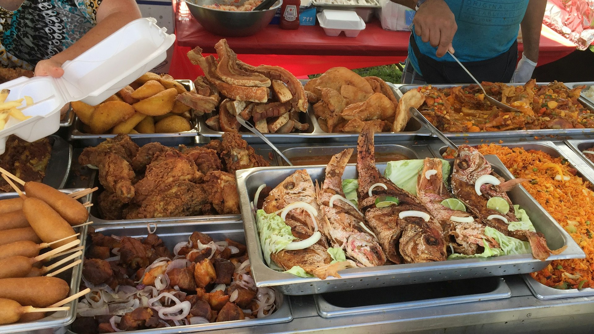 A street-food stand has many metal containers filled with different Latin foods such and empanadas and rice dishes