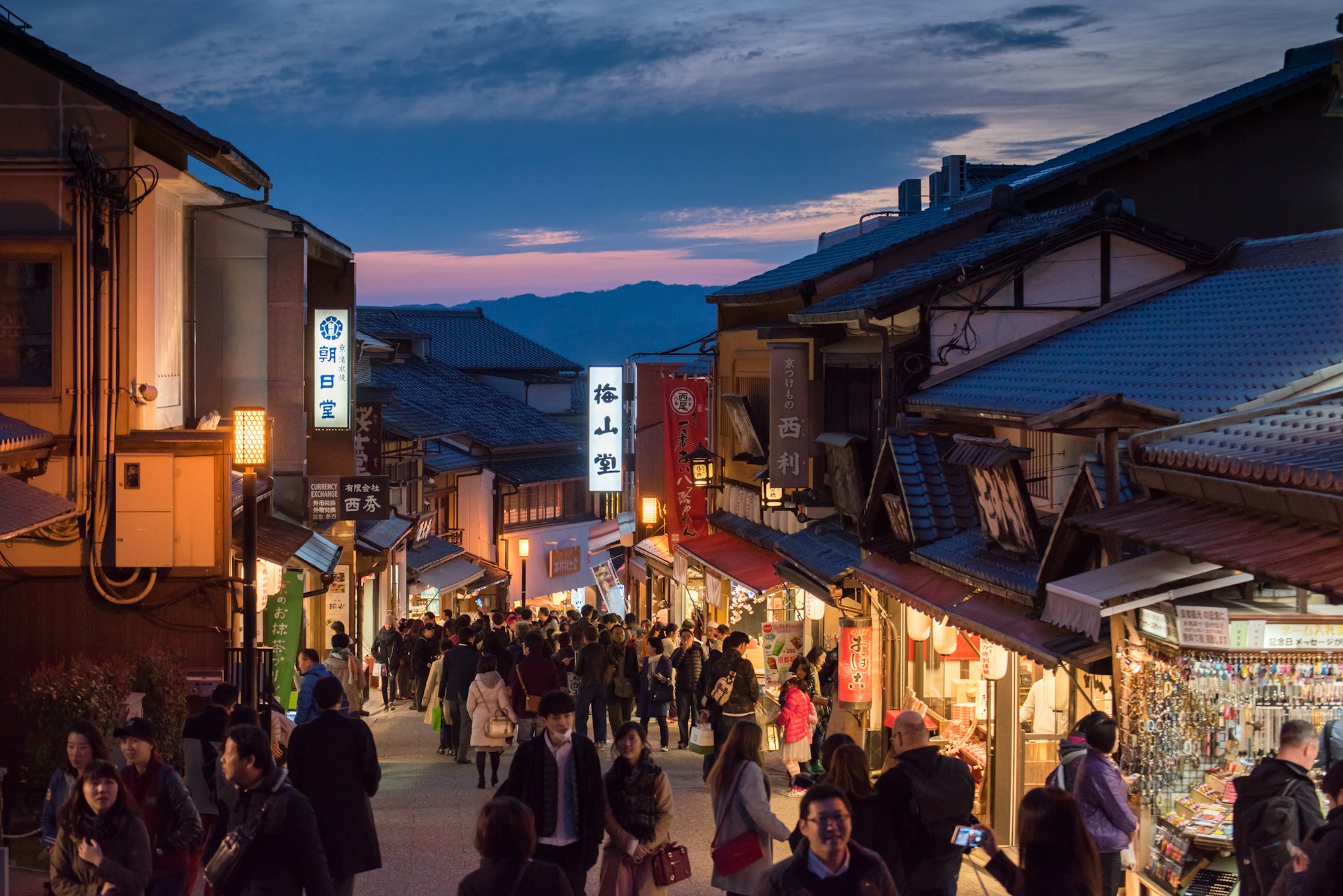 People walk down a steep street among shops with mountains in the distance, Gion district, Kyoto, Japan
