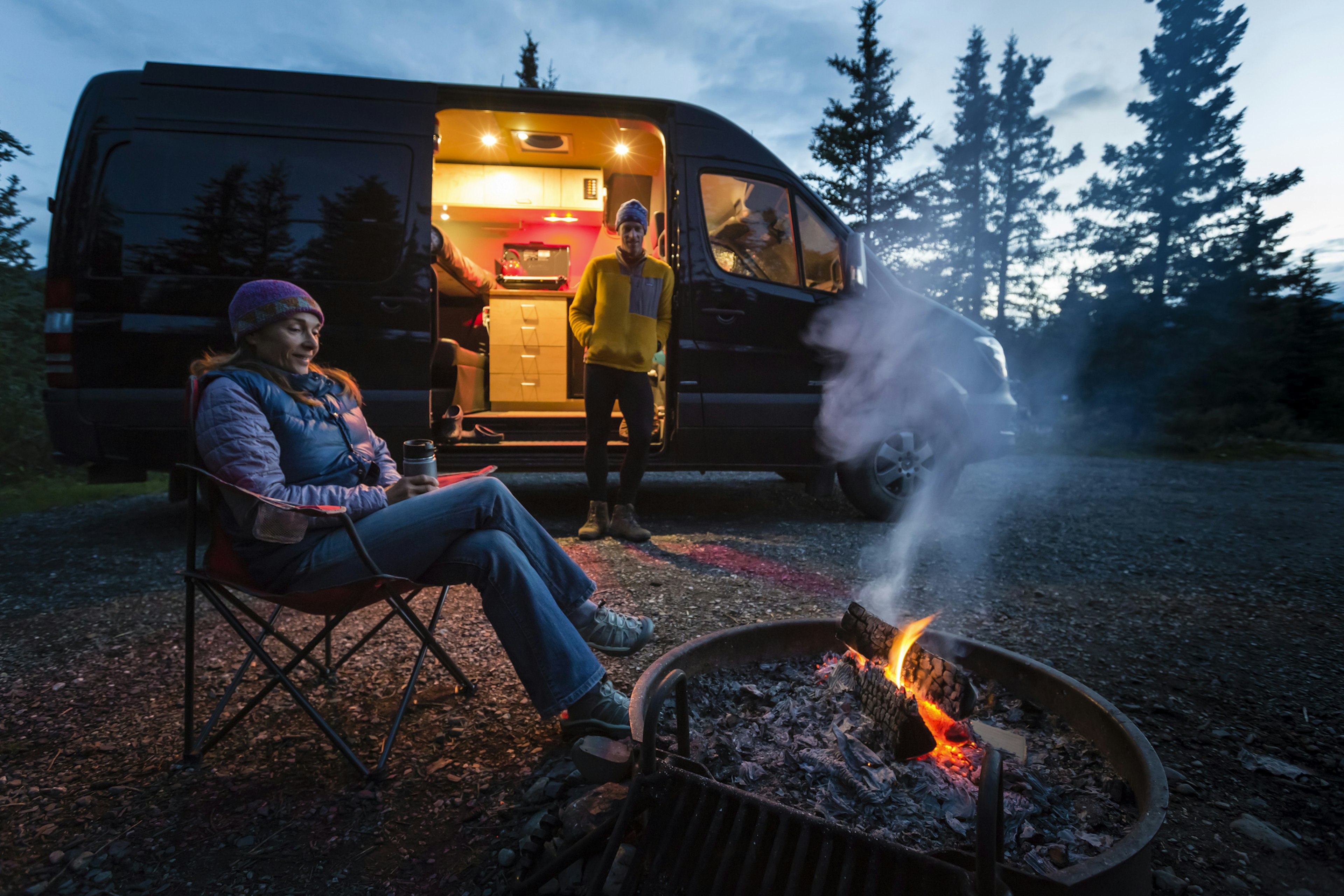 Two people sitting outside their camper van next to a fire at dusk in Denali National Park