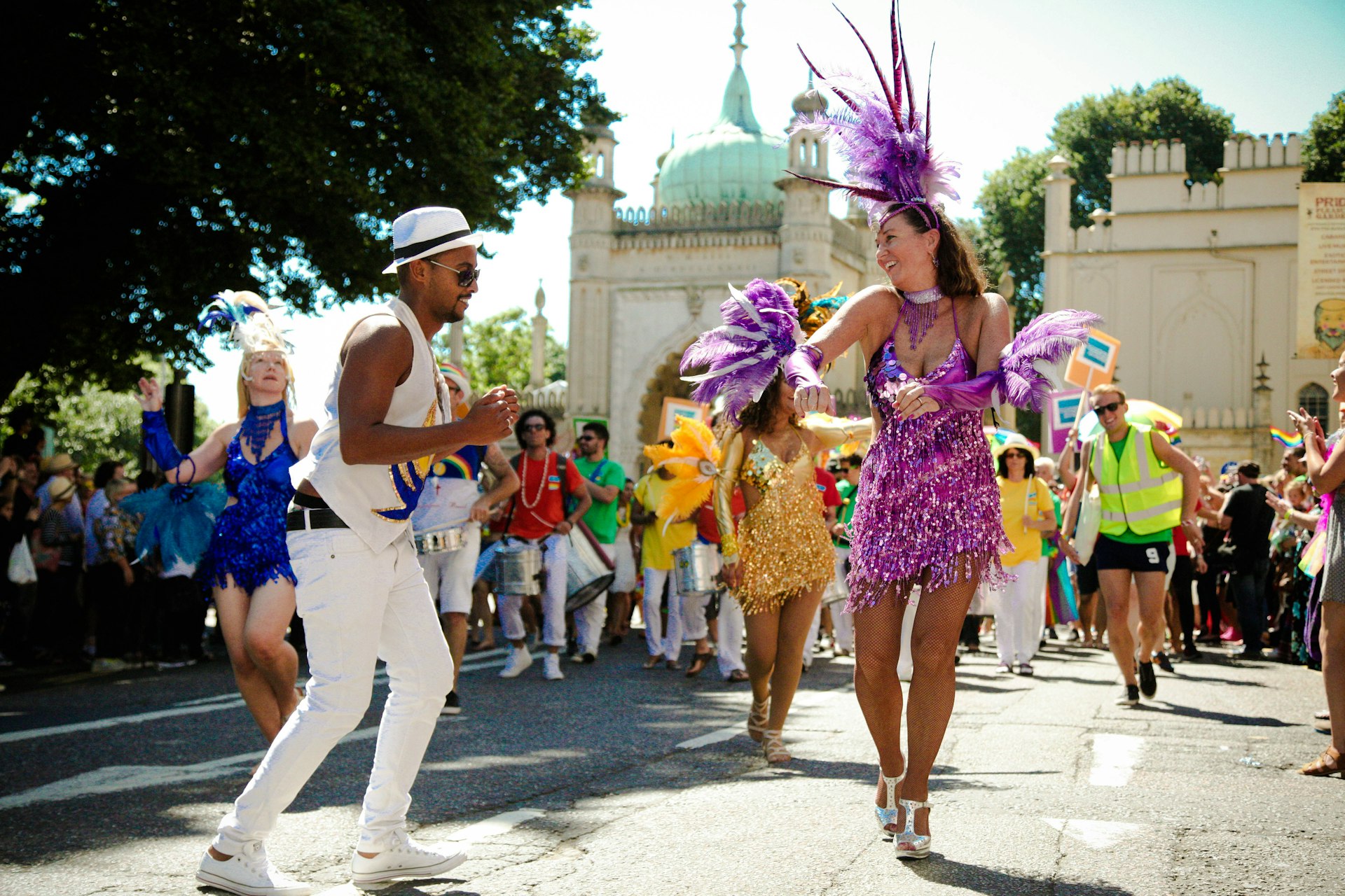 A large group of people in colorful outfits dance in the street outside the Royal Pavilion as part of Brighton's Pride parade