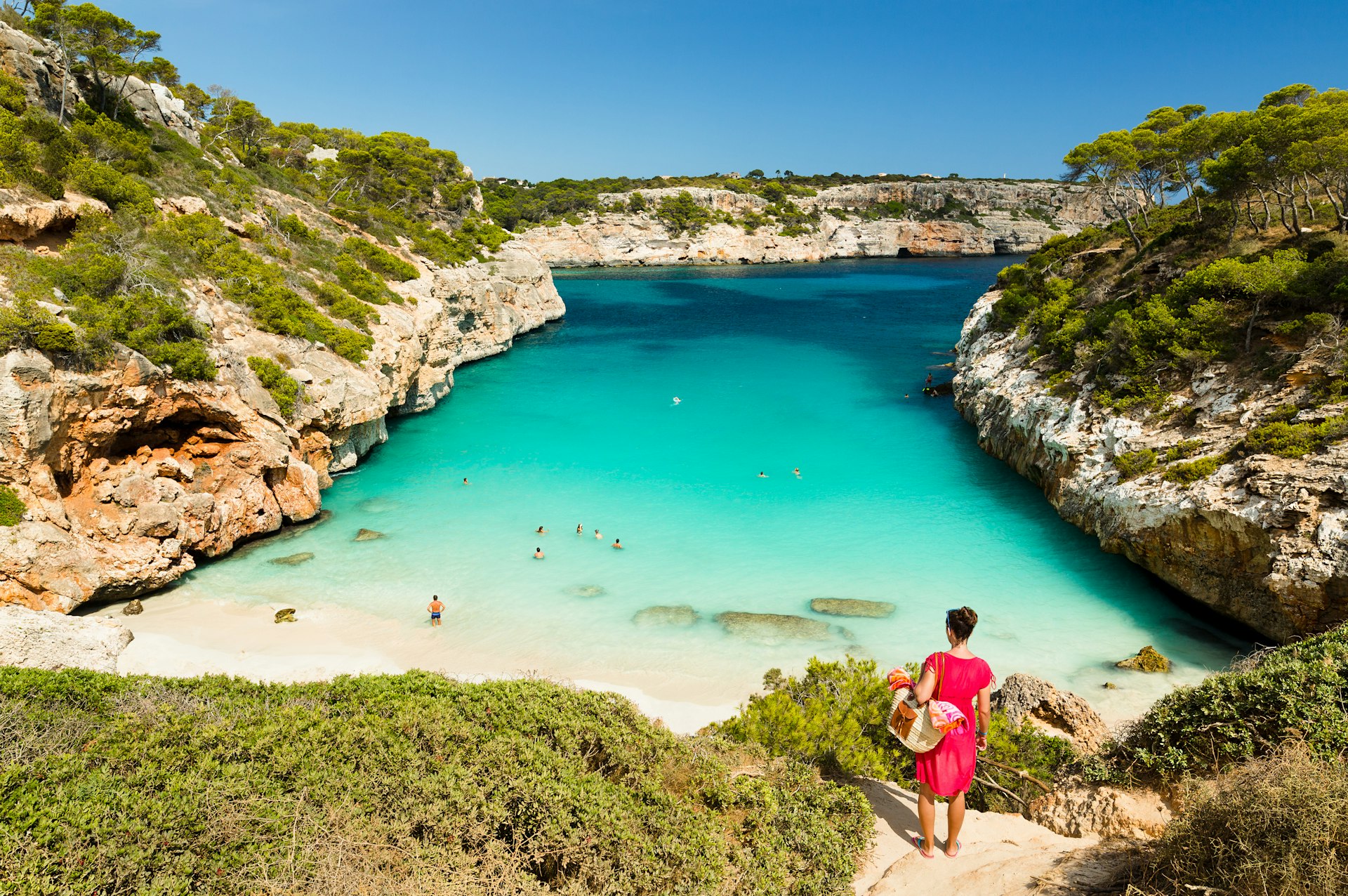A woman in a red cover-up stands looking over a stunning beach in Mallorca. 