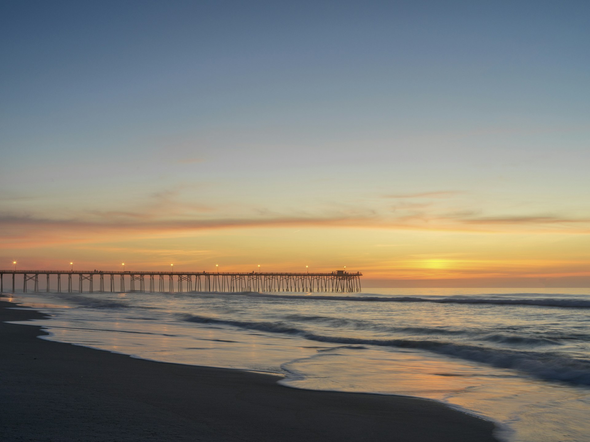 A wooden fishing pier stretches out into the ocean as the sun rises turning the sky orange