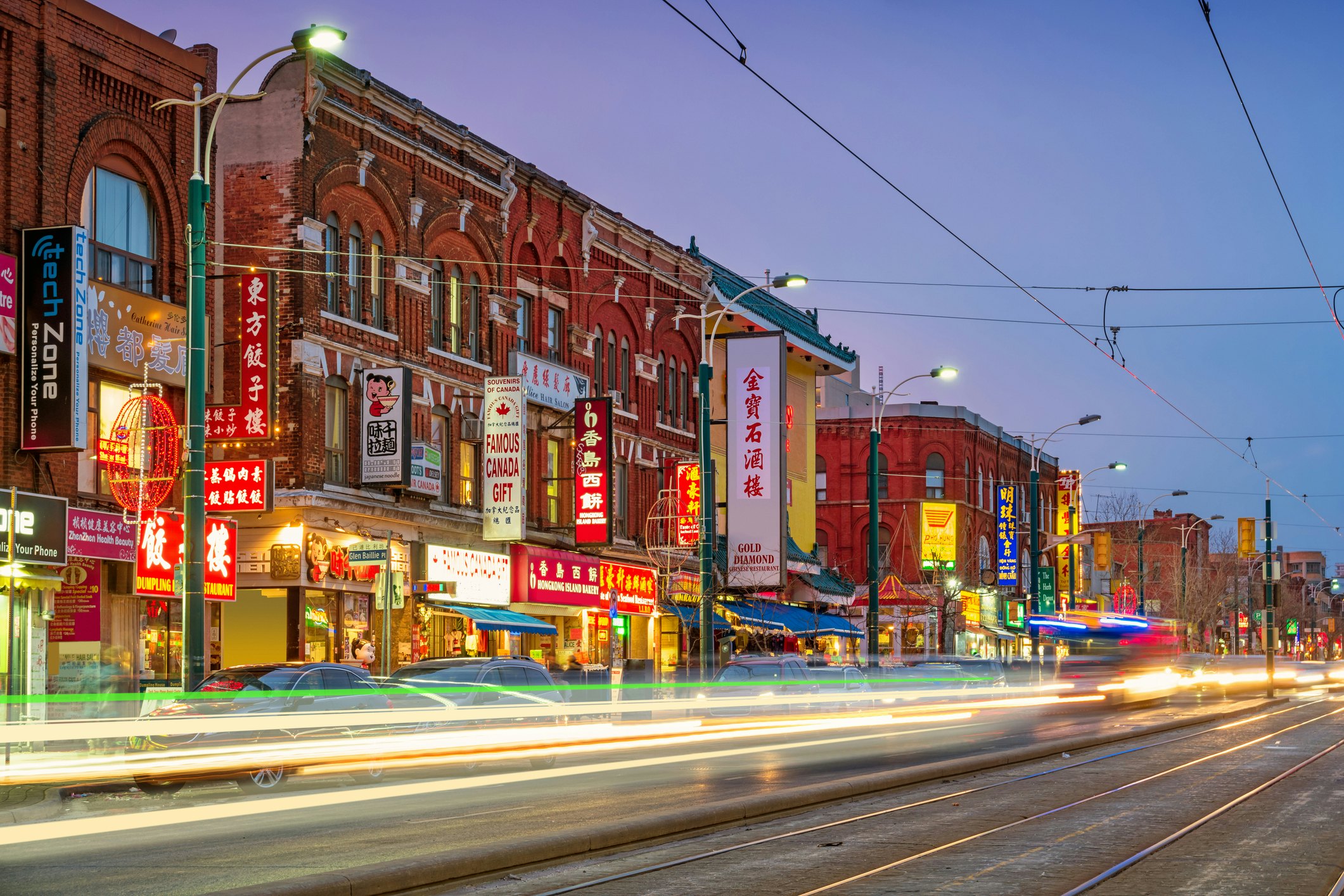 Lights blur as cars speed along the road outside a parade of shops with signs in English and Chinese