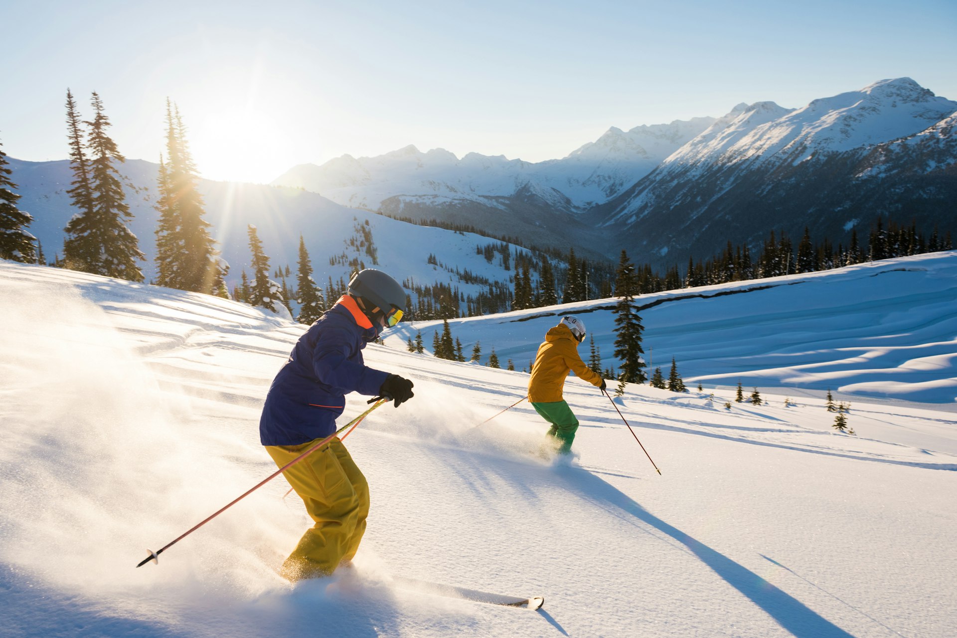 Skiers descend a powdery slope in a mountainous landscape bathed in bright sunshine