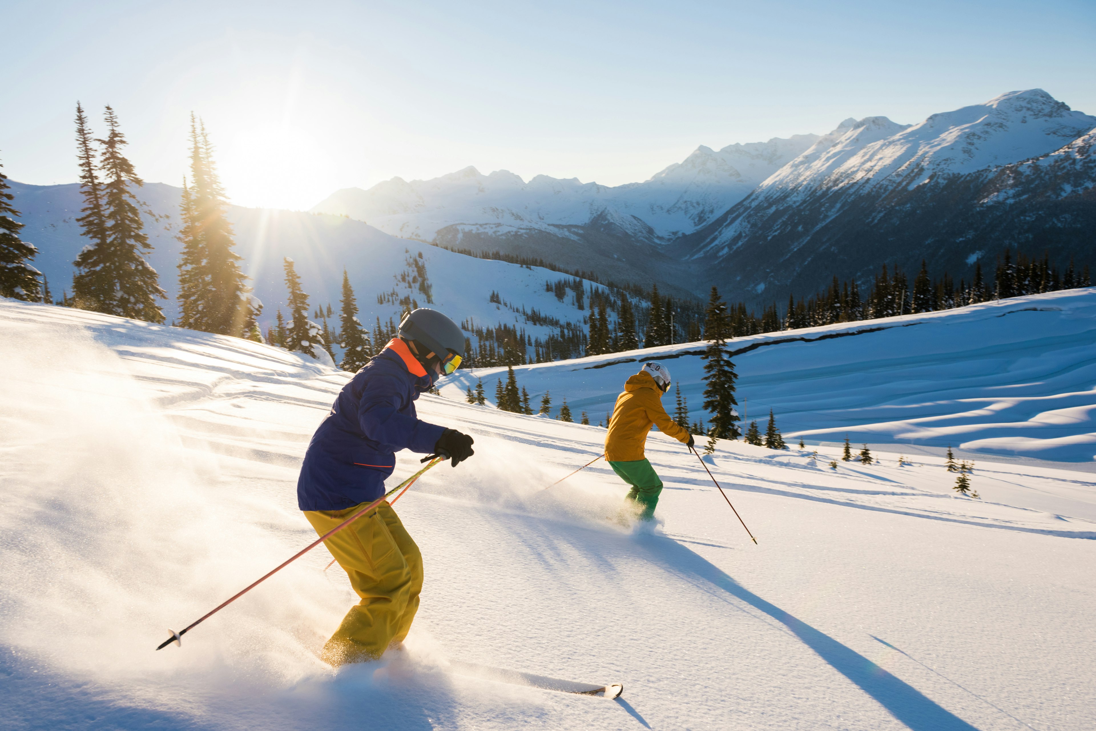 Skiers descend a powdery slope in a mountainous landscape bathed in bright sunshine