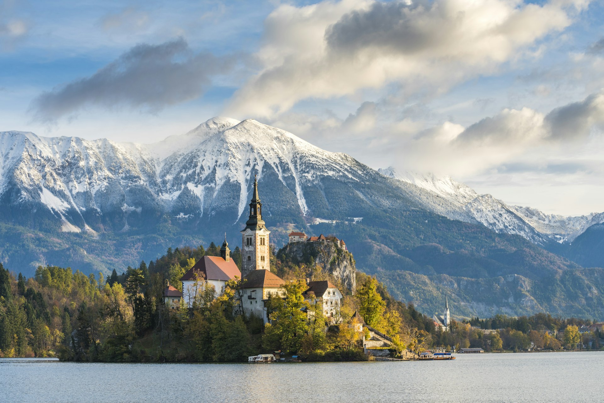 A beautiful church rises out of the trees in front of a lake with mountains in the background. 