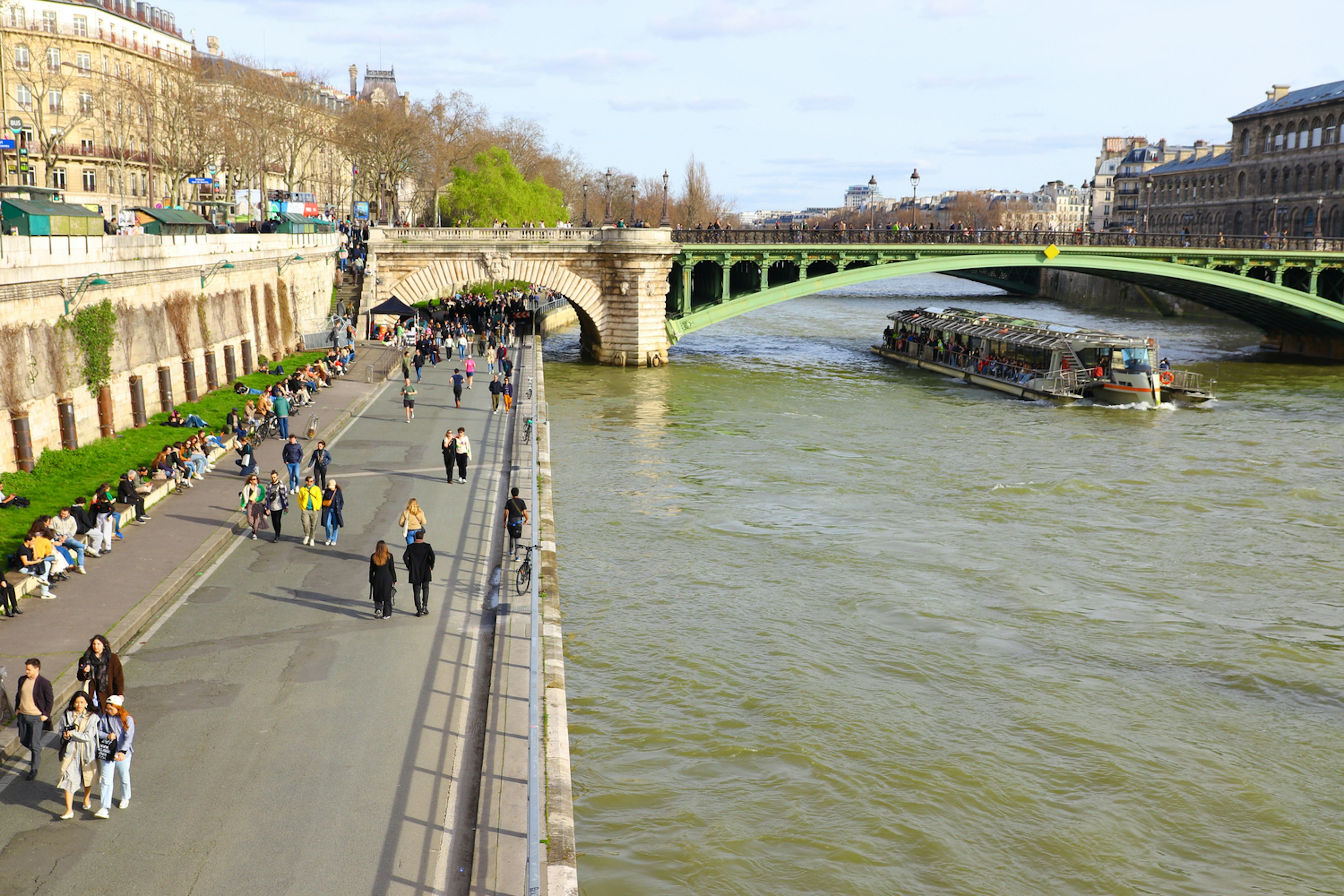 People follow a walkway beside a river heading towards a bridge