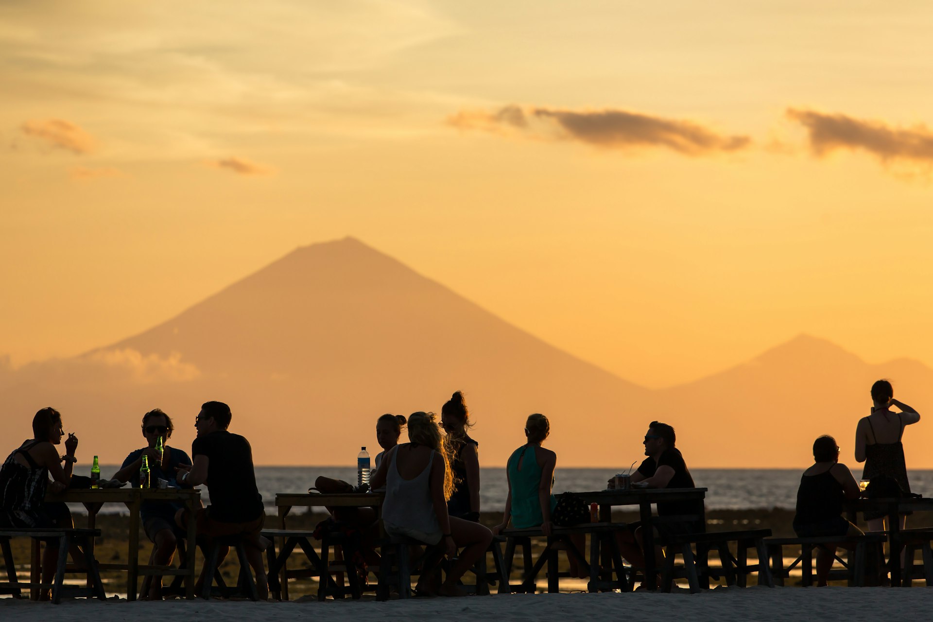 People at a beach bar are silhouetted as the sun sets behind a volcano