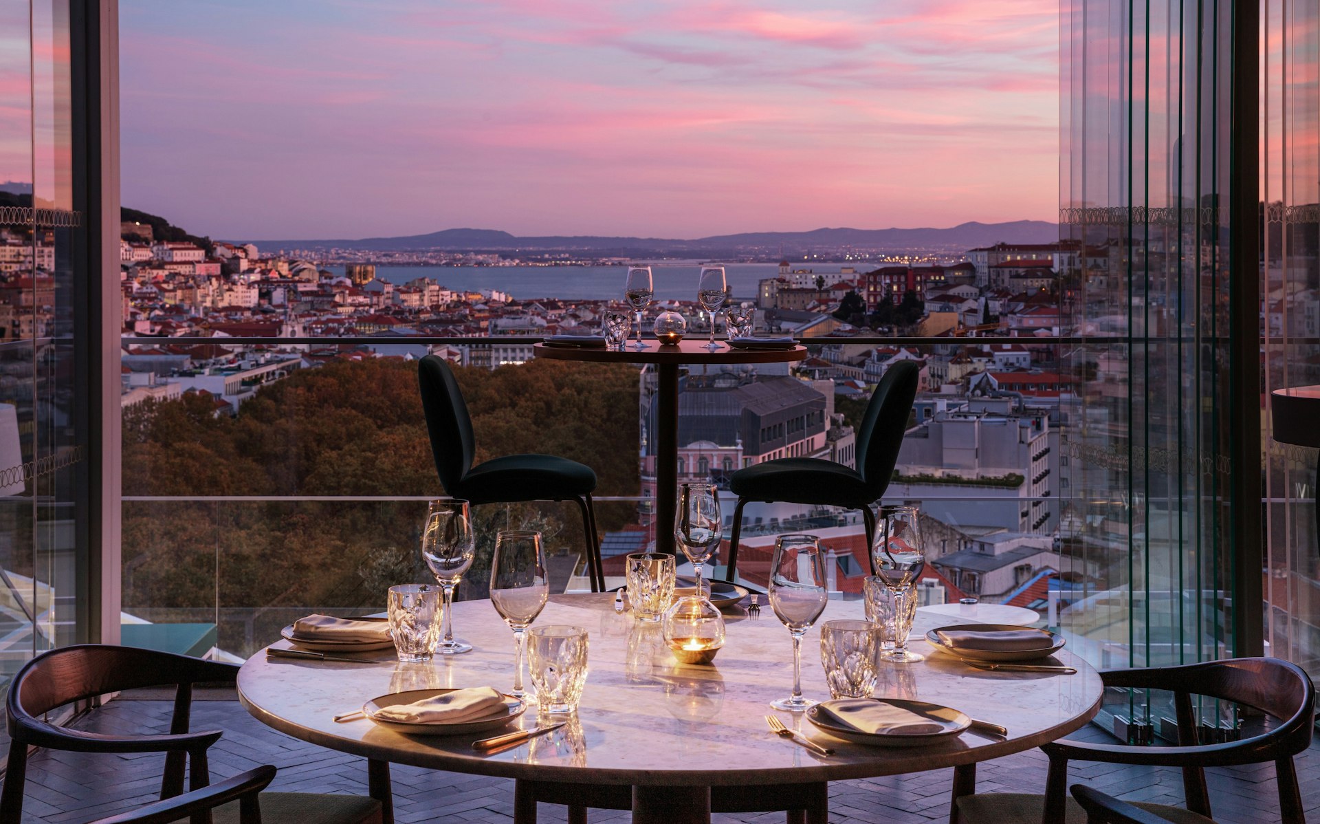 A table set next to a balcony with a view stretching over a city towards a river at dusk