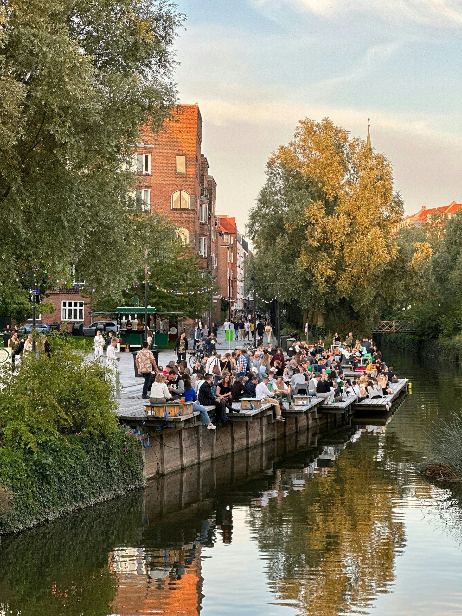 People are sat out on decking with drinks near a canal running through a town