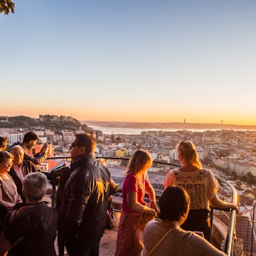 Tourists at Belvedere of Our Lady of the Hill viewpoint, looking at the cityscape of Lisbon at sunset.