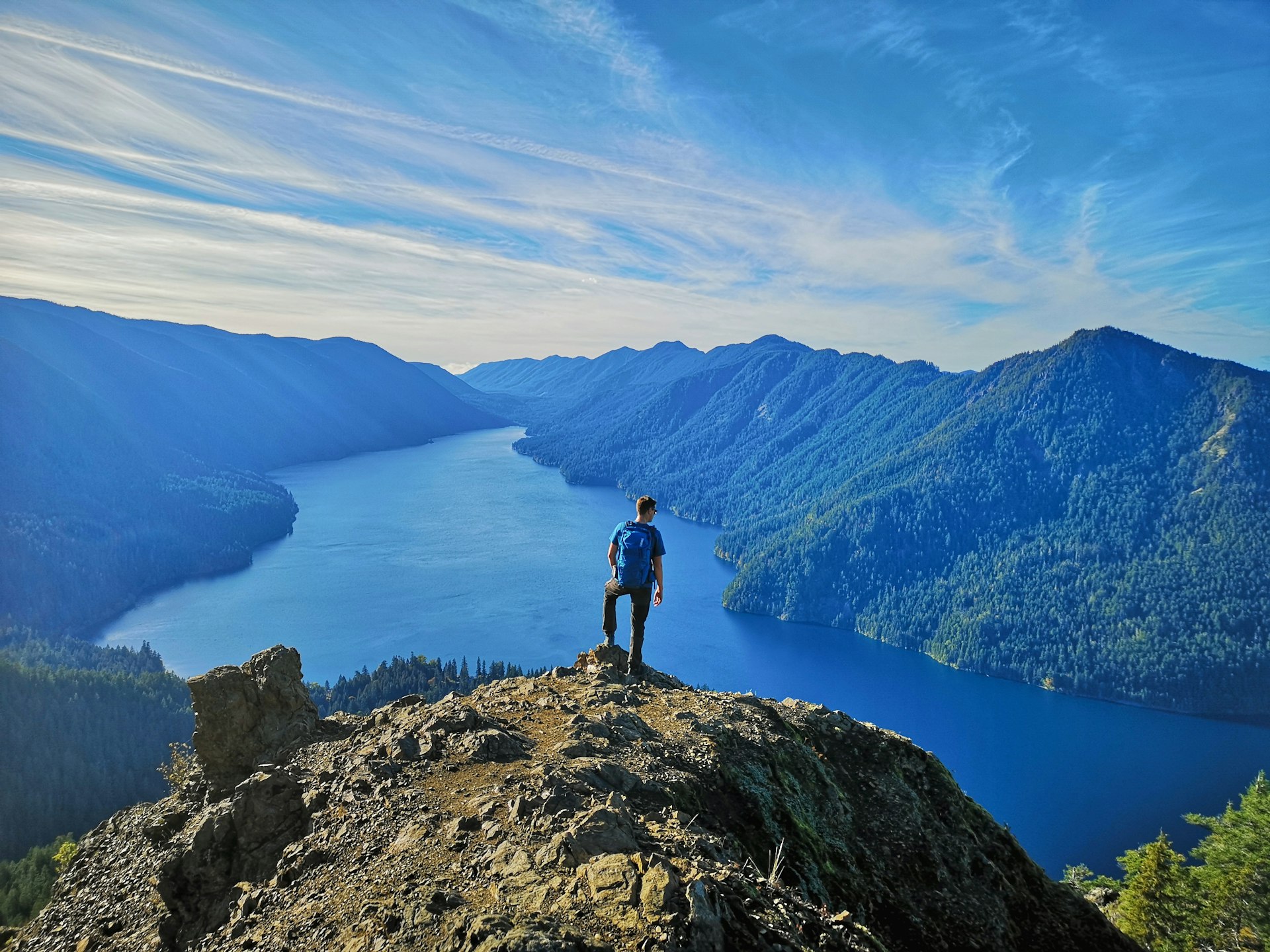 A man stands on a mountain peak looking down towards a lake